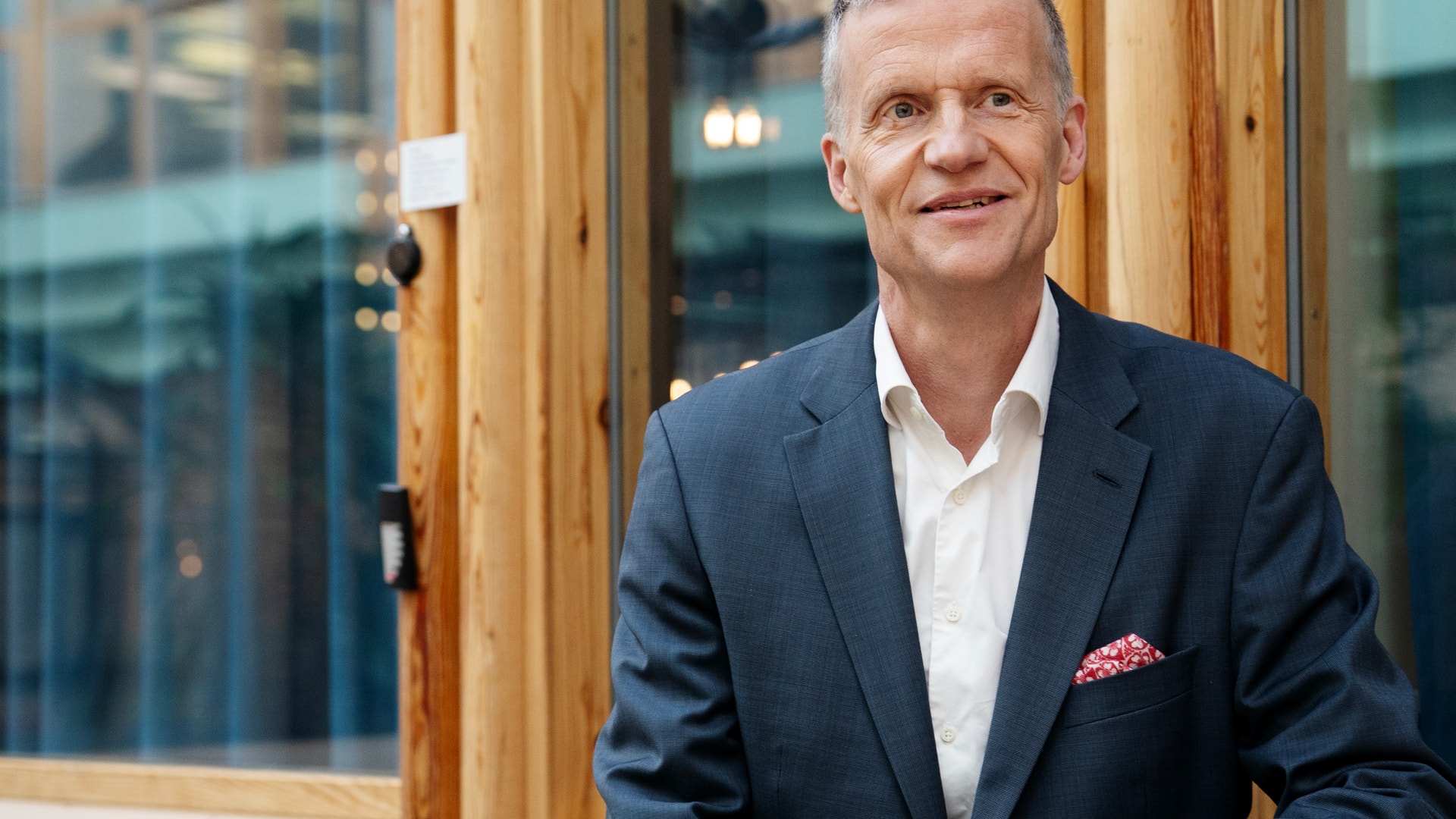 Porträtt av en man i mörkblå kostym och vit skjorta framför en trävägg med glasfönster i bakgrunden.