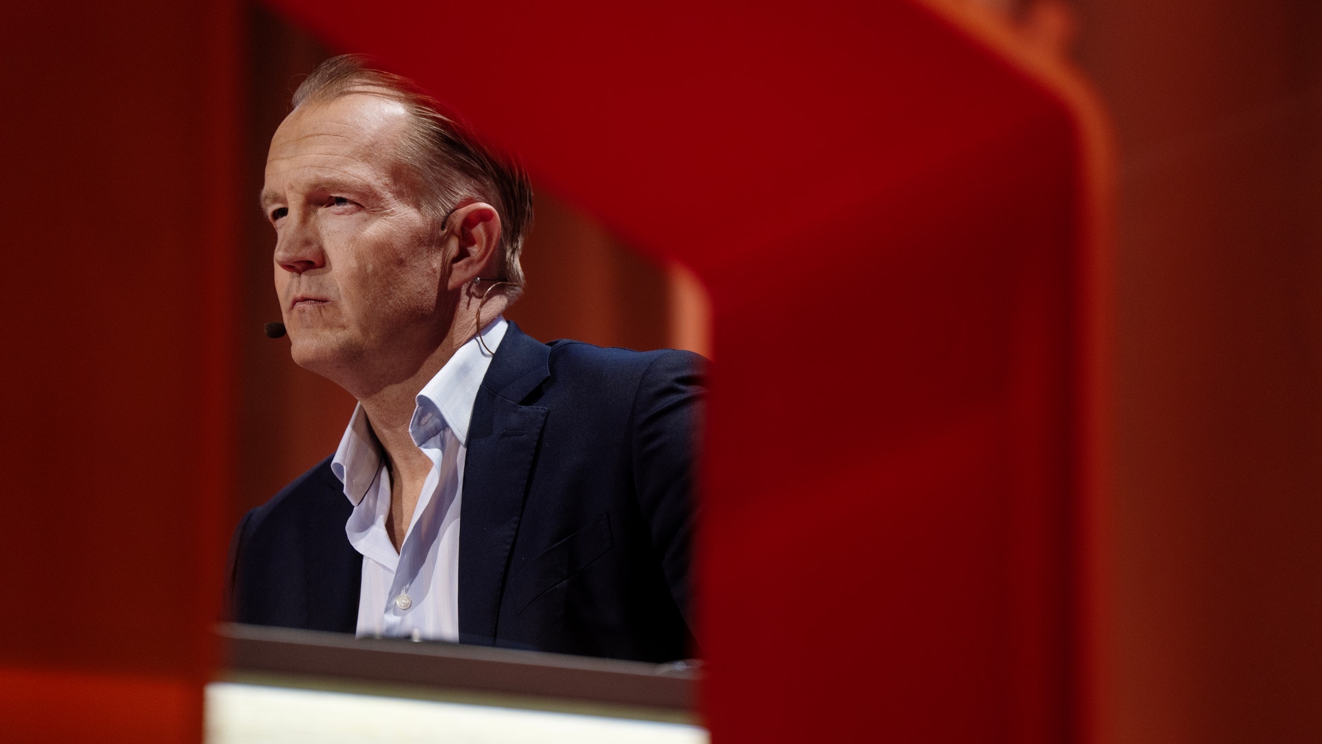 En man i mörk kostym talar vid ett podium framför en röd bakgrund.