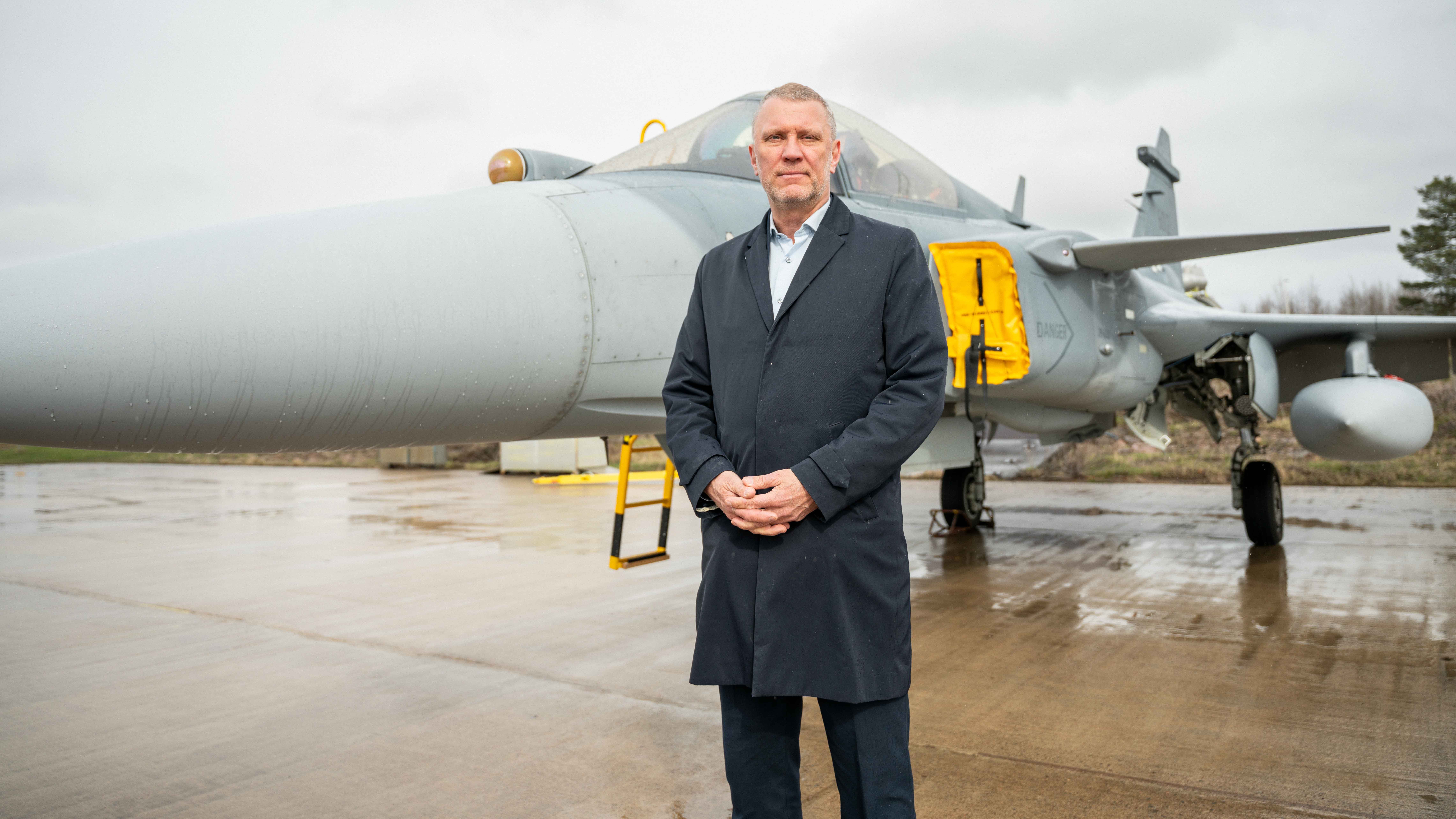 Man i mörkblå kappa står framför ett militärflygplan på en våt landningsbana.