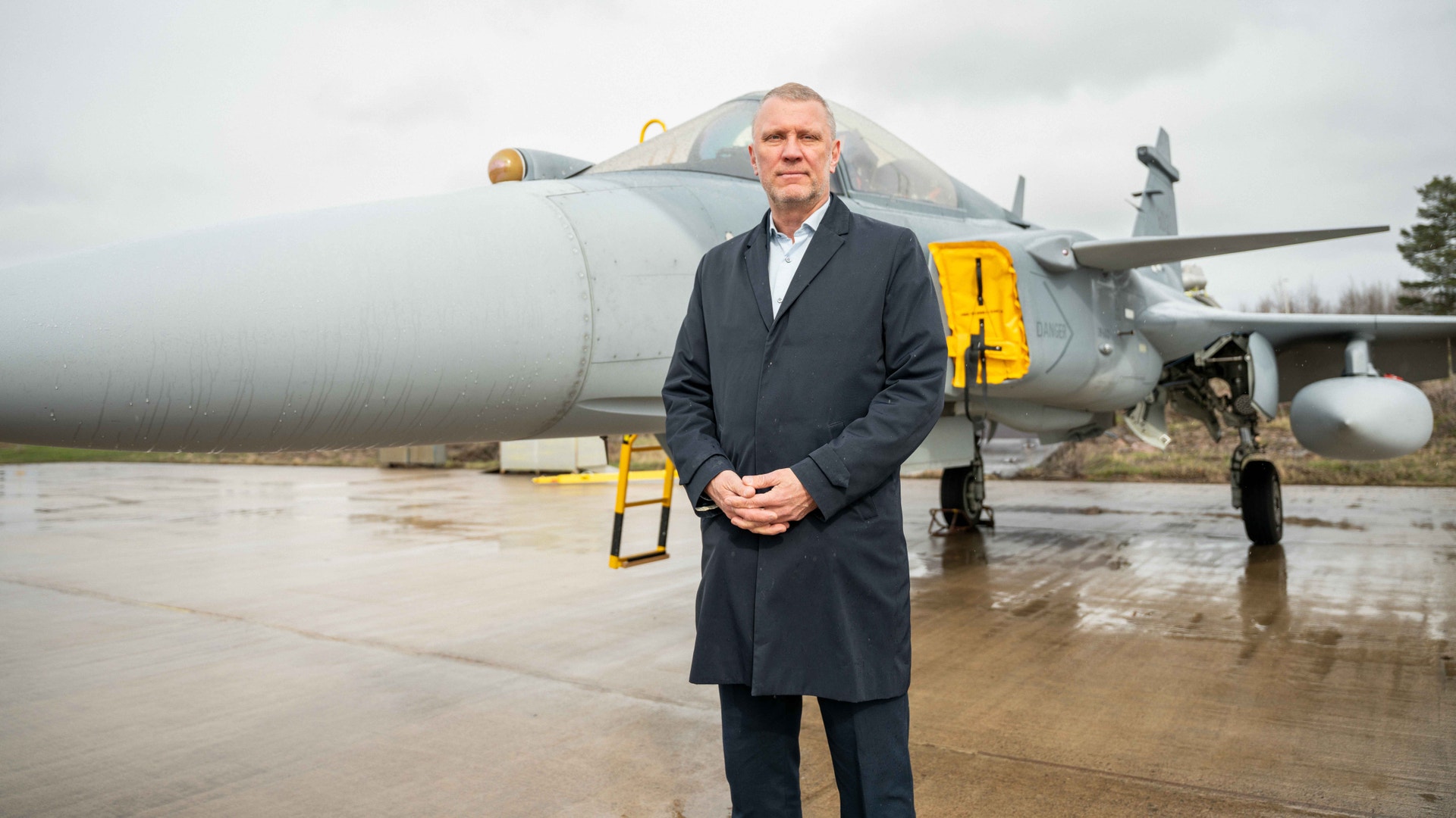 Man i mörkblå kappa står framför ett militärflygplan på en våt landningsbana.