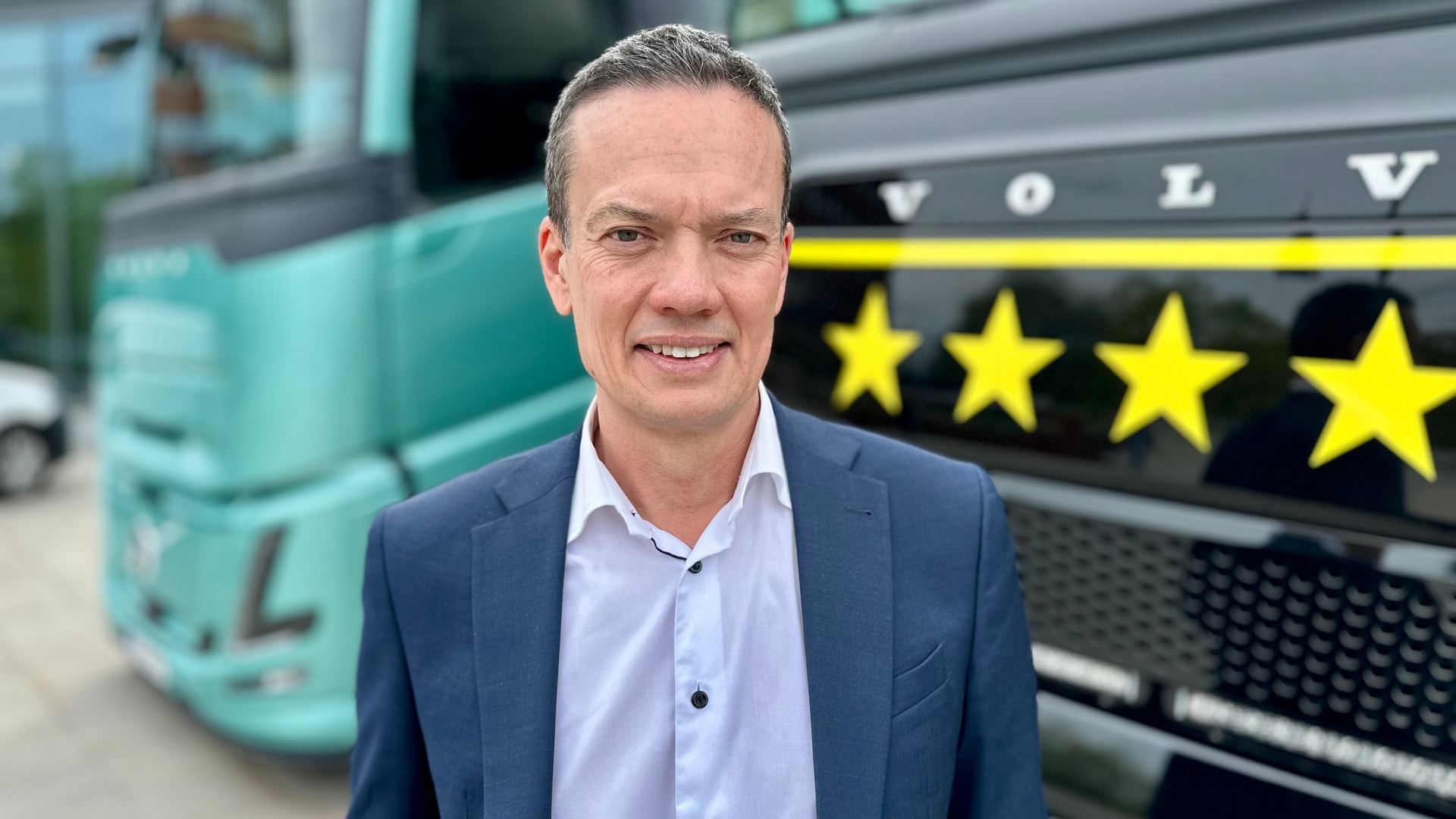 Man i blå kavaj och ljusblå skjorta står framför Volvo-lastbilar med gula stjärnor i bakgrunden.