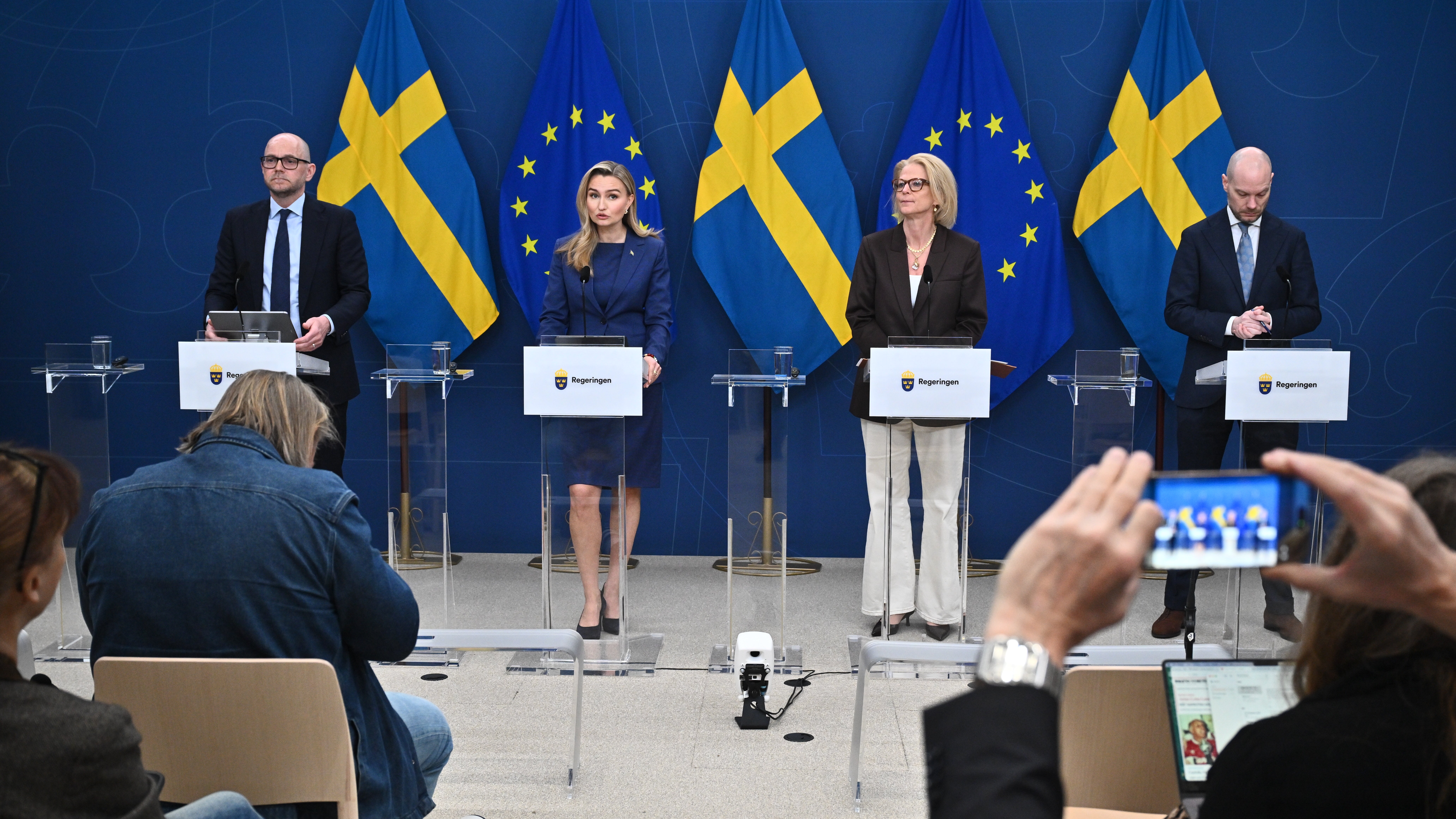 Fyra personer står vid podier under en blå bakgrund med svenska flaggor och EU-stjärnor. Journalister och publik sitter framför scenen.