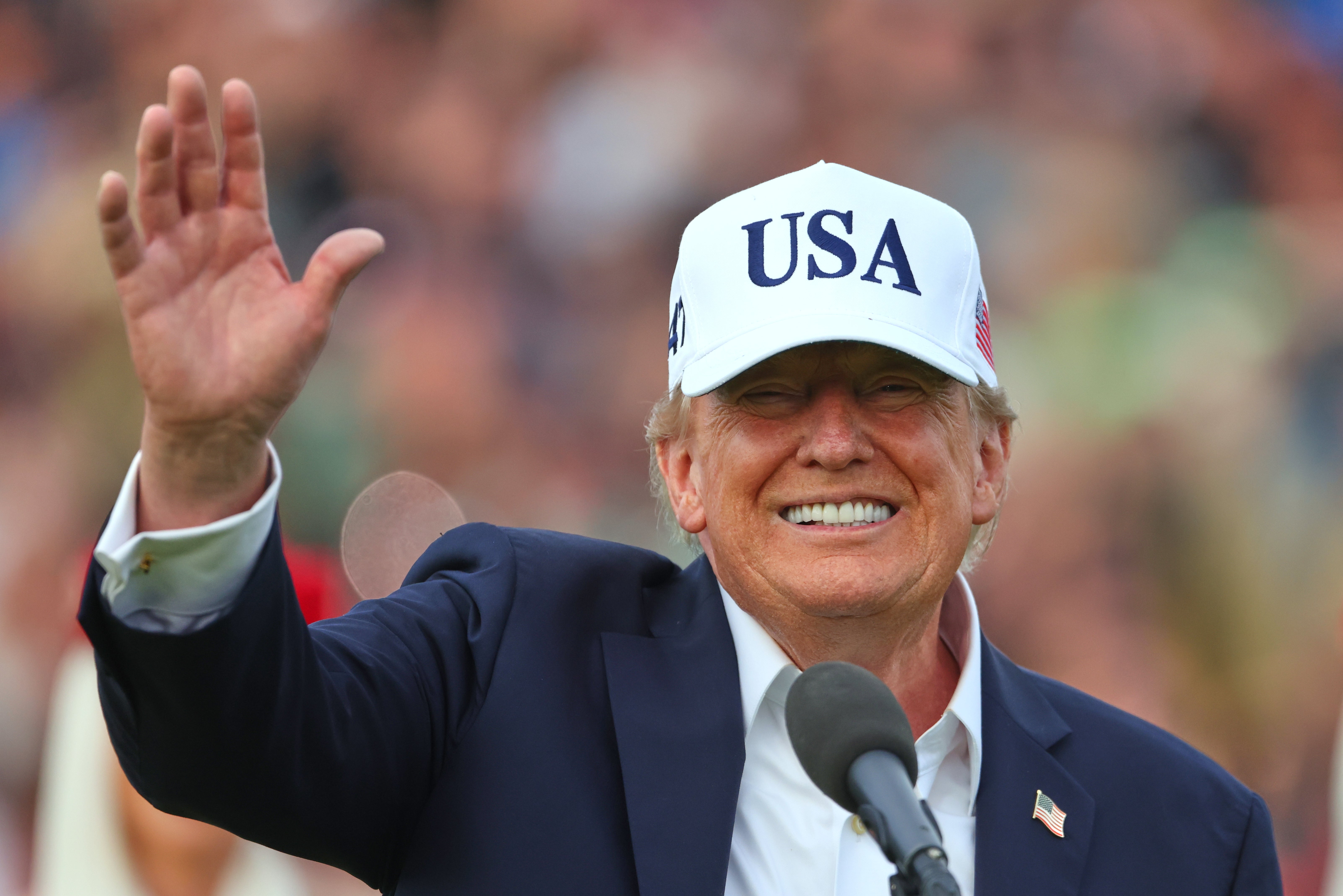 Donald Trump ler och vinkar framför en mikrofon, iklädd kavaj och en vit keps med texten USA.
