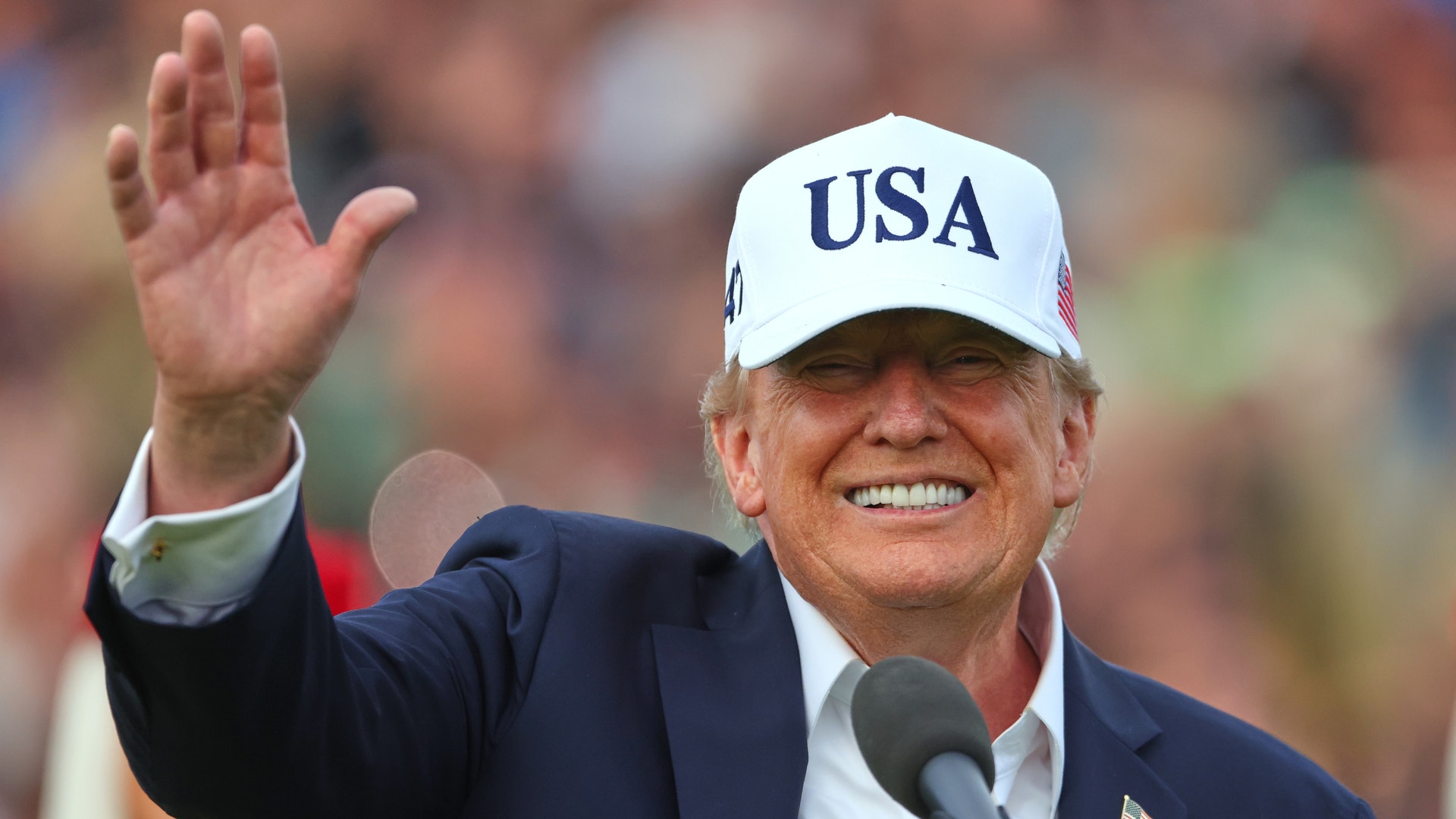 Donald Trump ler och vinkar framför en mikrofon, iklädd kavaj och en vit keps med texten USA.