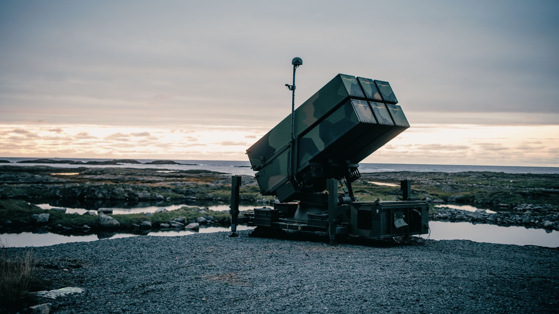 Ett militärt luftförsvarsradarsystem monterat på en upphöjd position med utsikt över ett landskap med vatten och ängar.