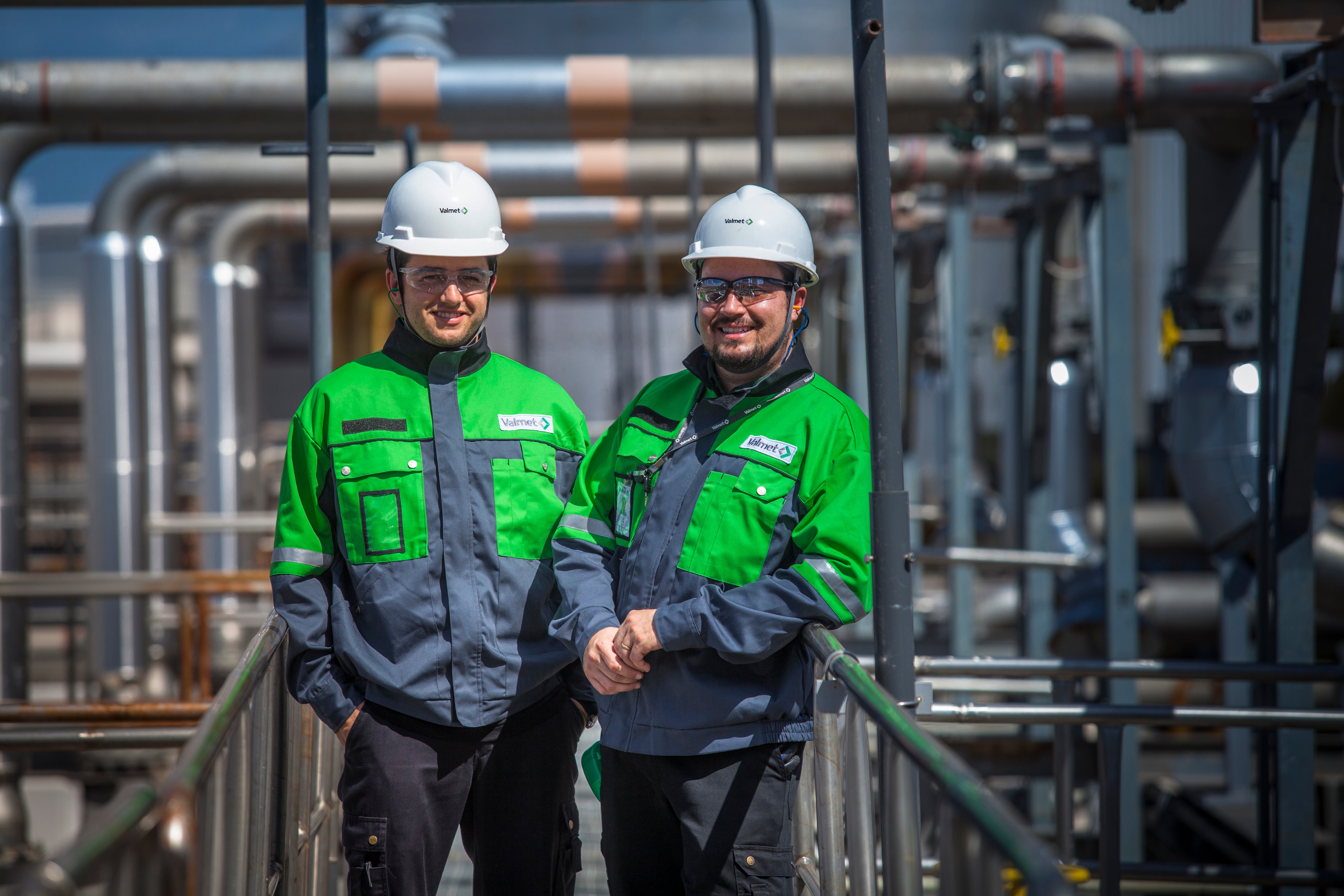 Två arbetare i säkerhetsutrustning står på en metallplattform framför industriell rörledning och maskiner.