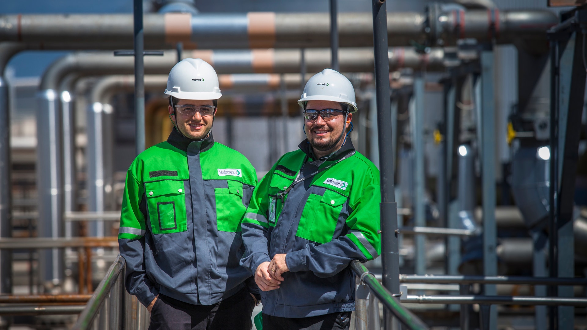 Två arbetare i säkerhetsutrustning står på en metallplattform framför industriell rörledning och maskiner.