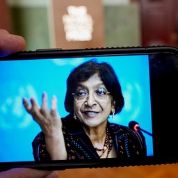 En hand håller en mobiltelefon som visar en leende kvinna med glasögon som talar i en mikrofon och gestikulerar. I bakgrunden syns en suddig sal med en talarstol.