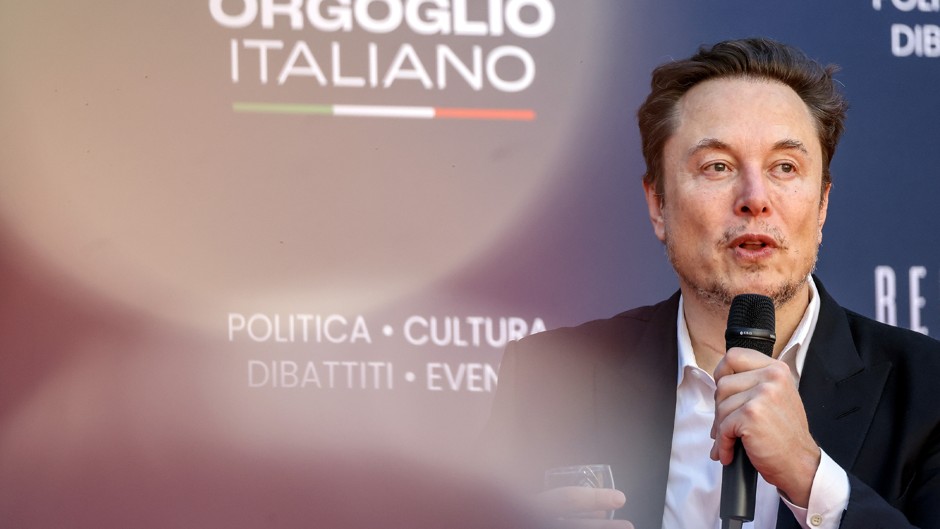 Man i mörk kostym talar i mikrofon vid en offentlig event med italiensk text i bakgrunden.