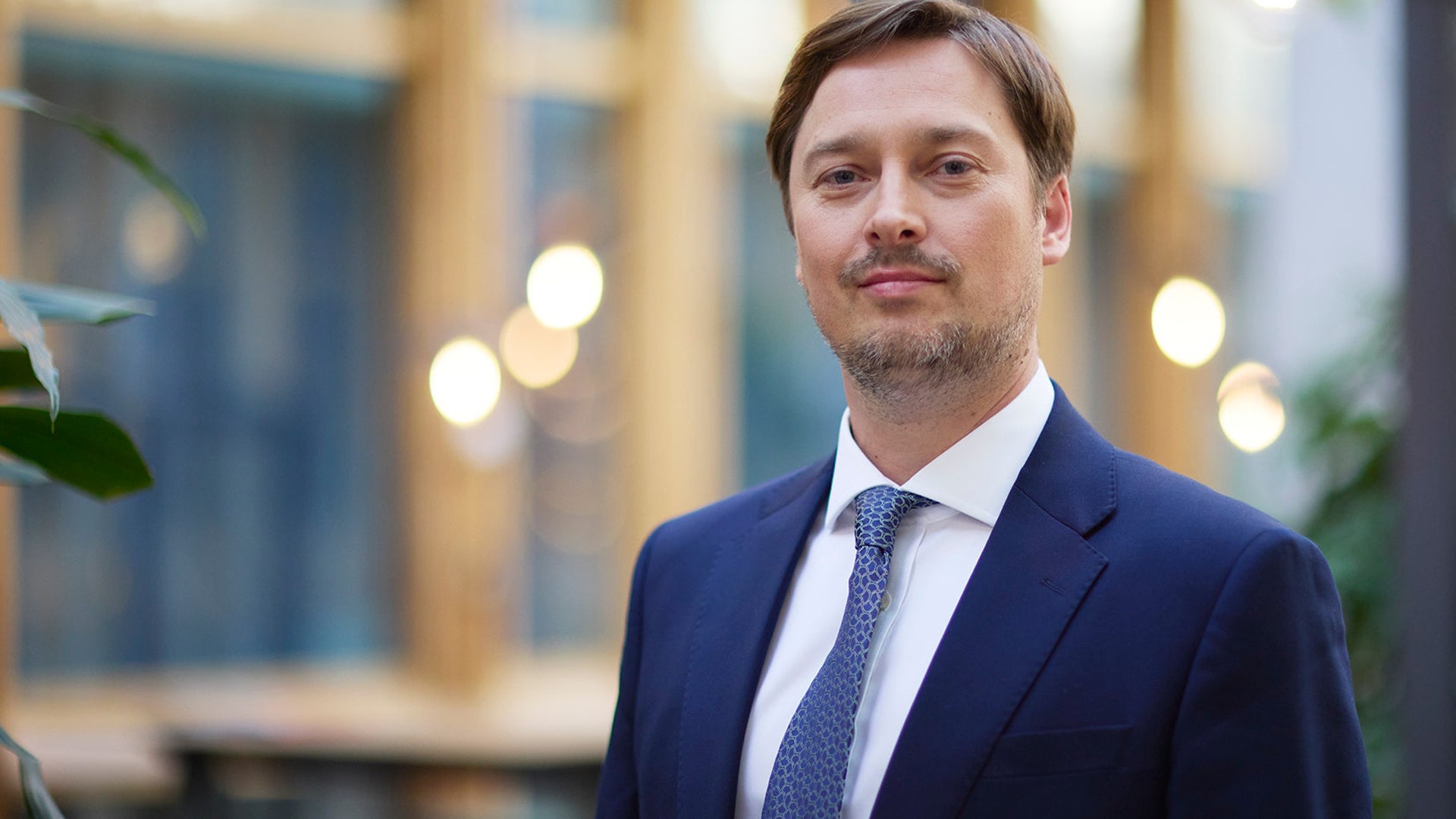 En man i mörkblå kostym och blå slips står framför en suddig bakgrund med varma ljus i vad som verkar vara en professionell miljö.