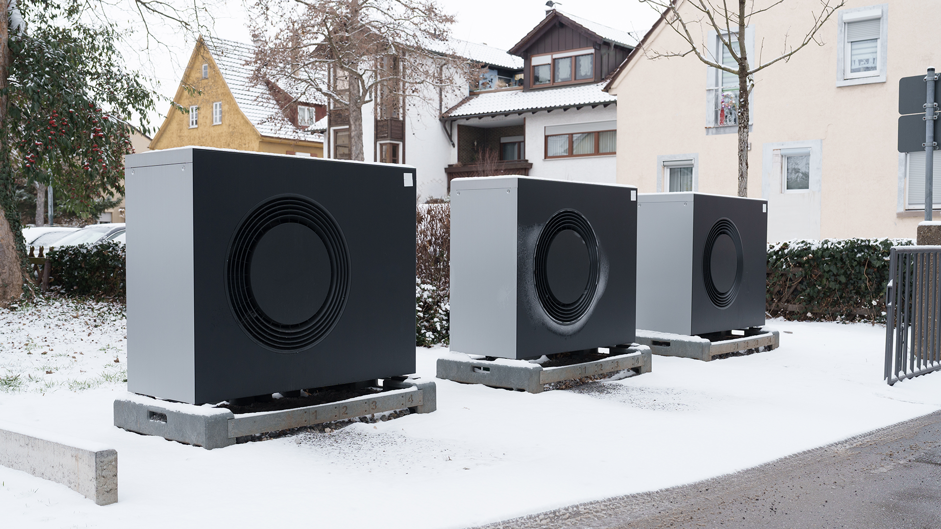 Tre vita värmepumpsenheter med svarta fläktar placerade på en snötäckt yta framför bostadshus.