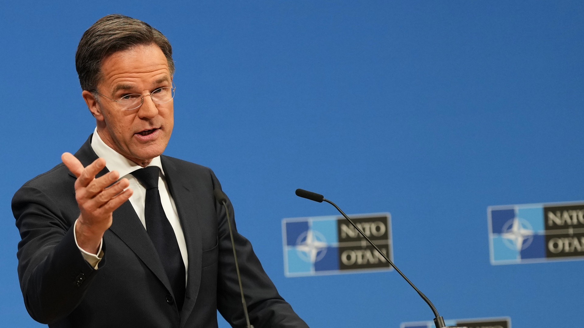 Man i kostym talar vid ett podium med NATO-loggor i bakgrunden.