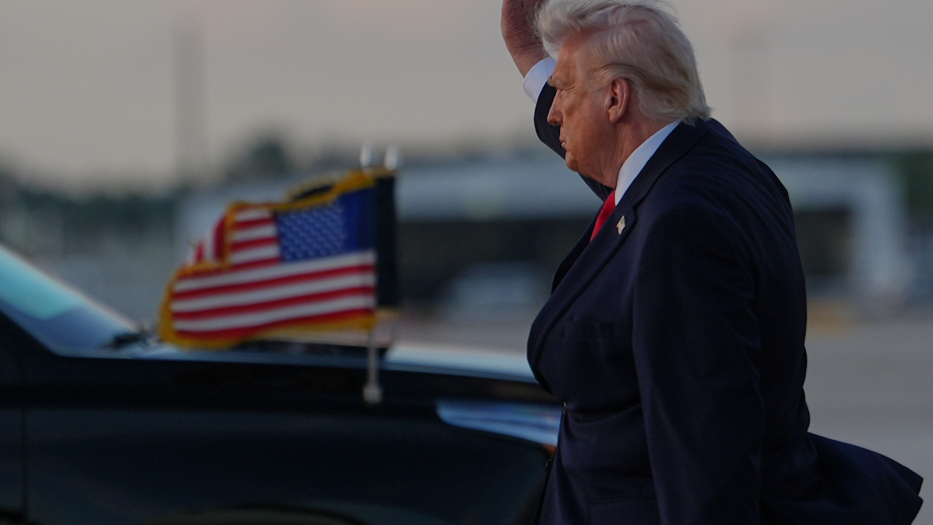 Man i mörk kostym och röd slips står med upphöjd hand framför amerikansk flagga.