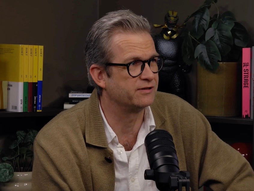En man med glasögon och beige kofta talar i en mikrofon under en podcast- eller intervjusession.