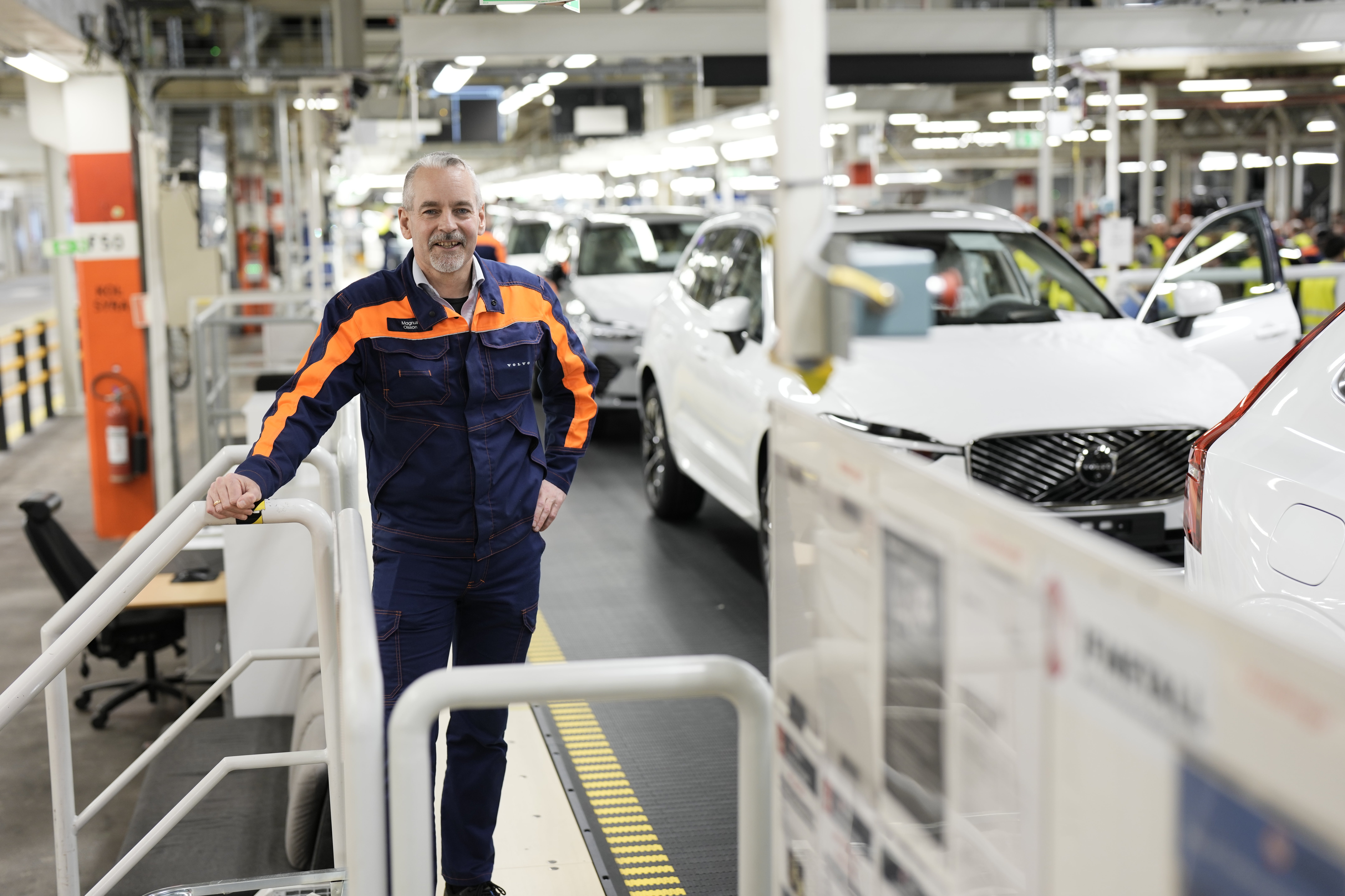 En arbetare i blå och orange arbetsoverall står i en bilfabrik omgiven av vita bilar under montering.