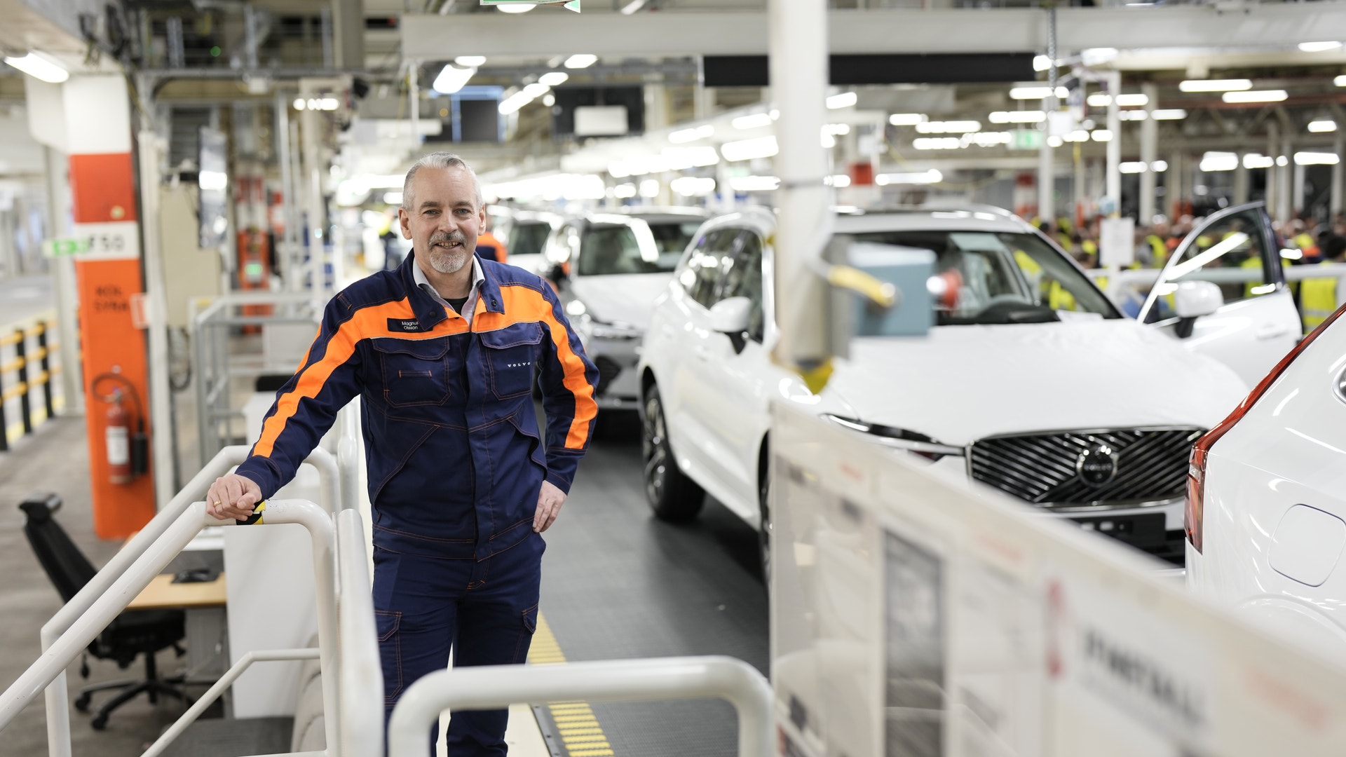 En arbetare i blå och orange arbetsoverall står i en bilfabrik omgiven av vita bilar under montering.