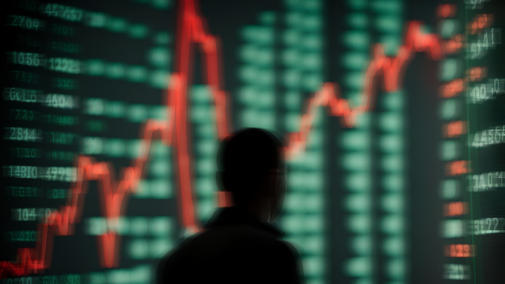 Silhuett av en person framför en aktiemarknadsskärm med röda och gröna kursgrafer och siffror.