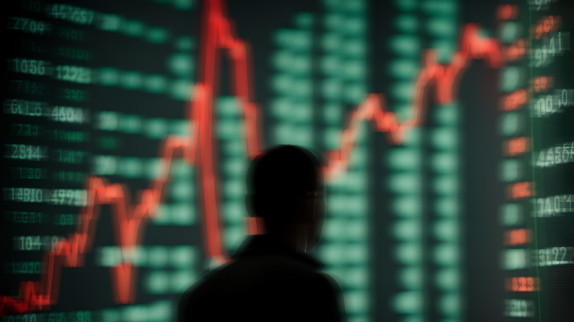 Silhuett av en person framför en aktiemarknadsskärm med röda och gröna kursgrafer och siffror.
