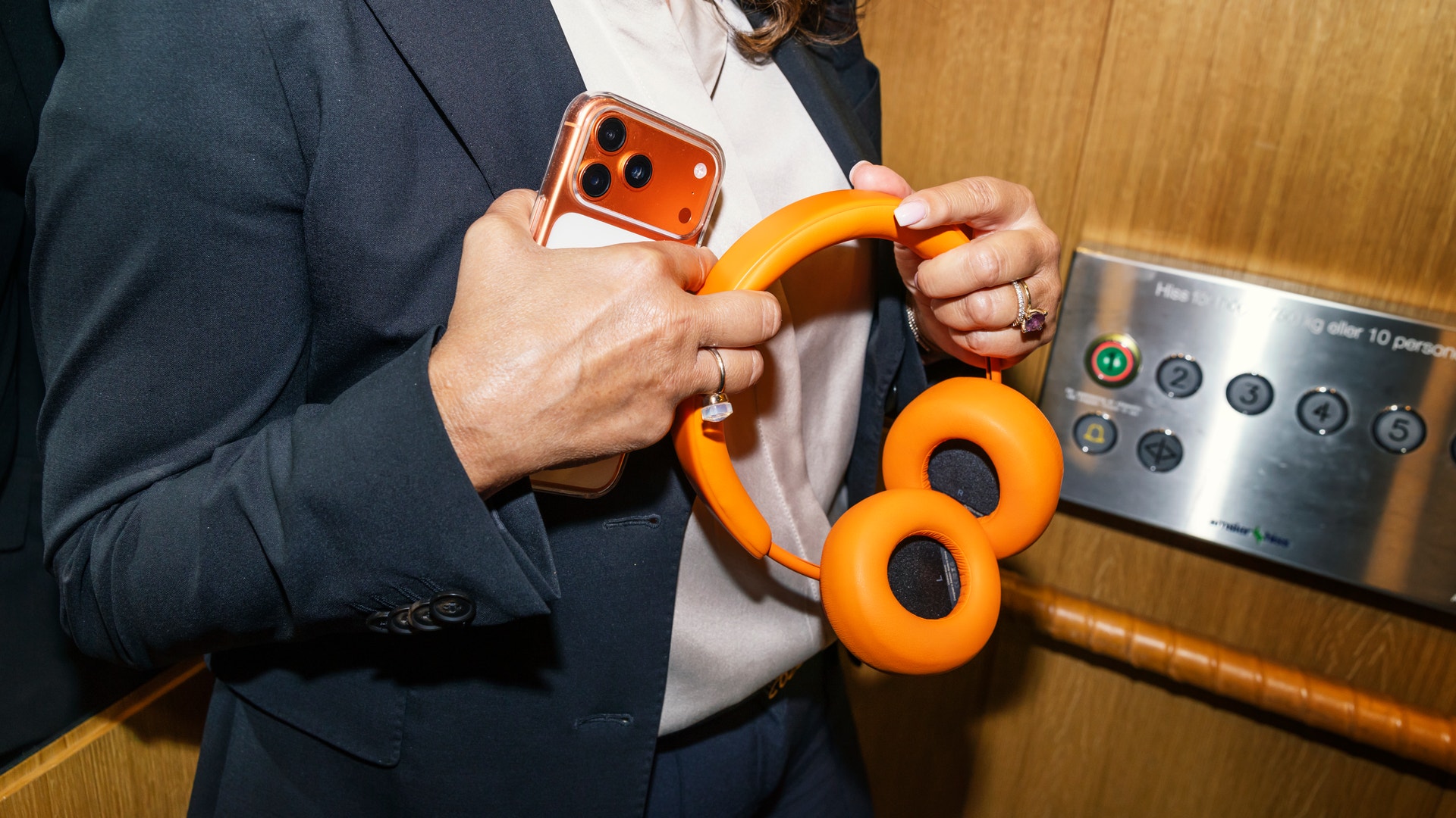 Person i mörkblå kavaj håller upp en orange mobiltelefonhållare och orange hörlurar framför sitt bröst.