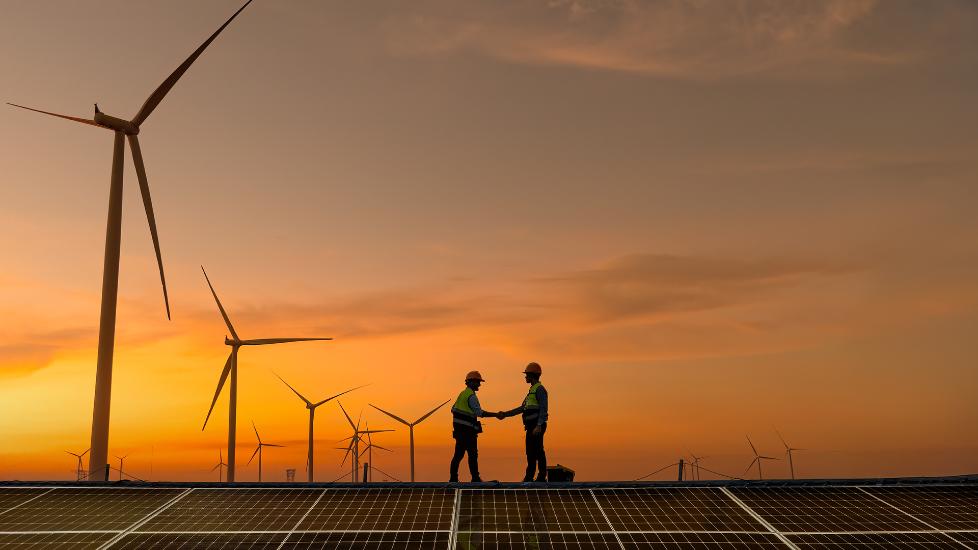 Två personer står på solpaneler och skakar hand med vindkraftverk i bakgrunden vid solnedgång, symboliserande förnybar energi och hållbar samarbete.