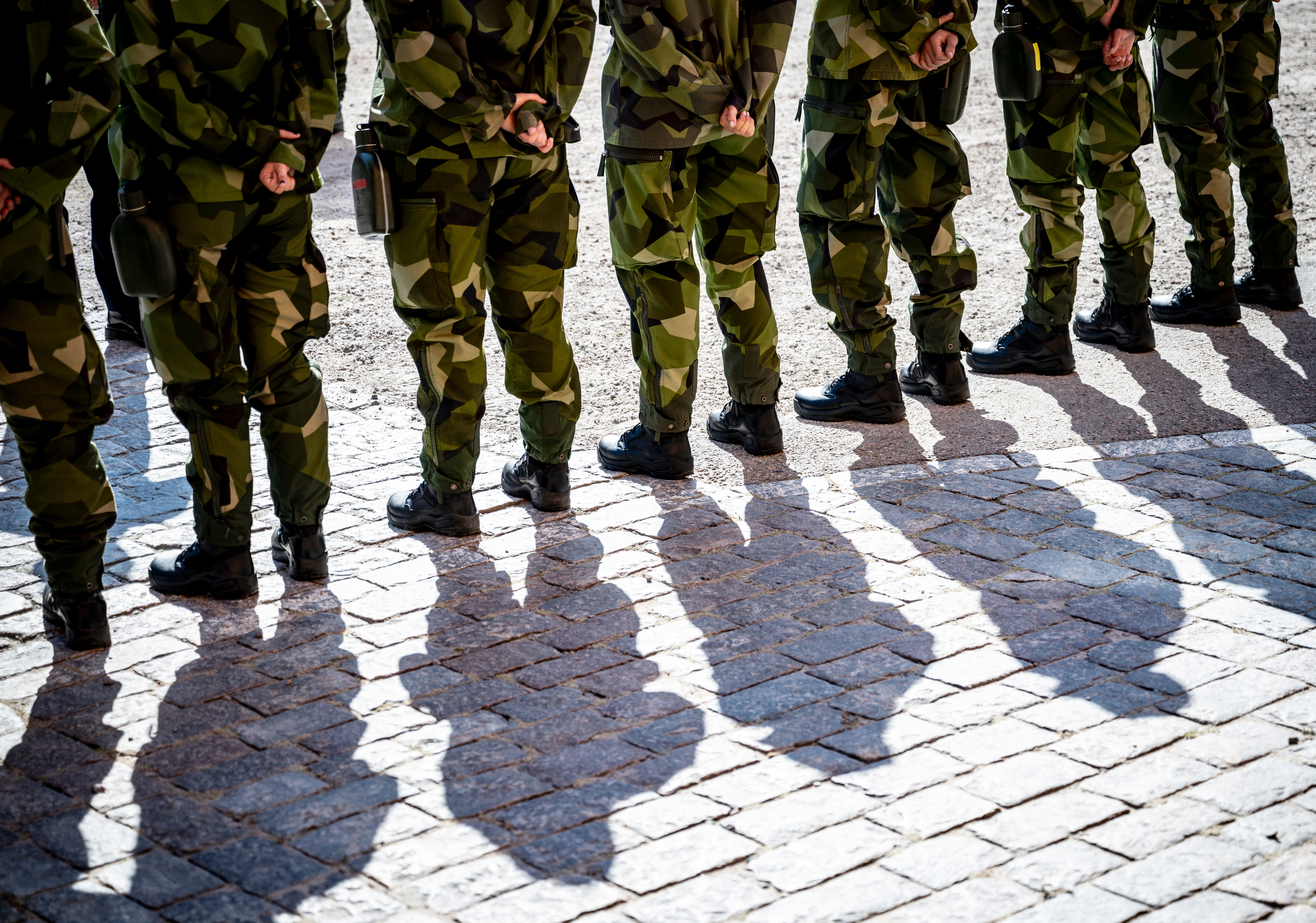 Soldater i kamouflageuniform står på rad på en stenlagd plats. Deras skuggor sträcker sig över stenarna.