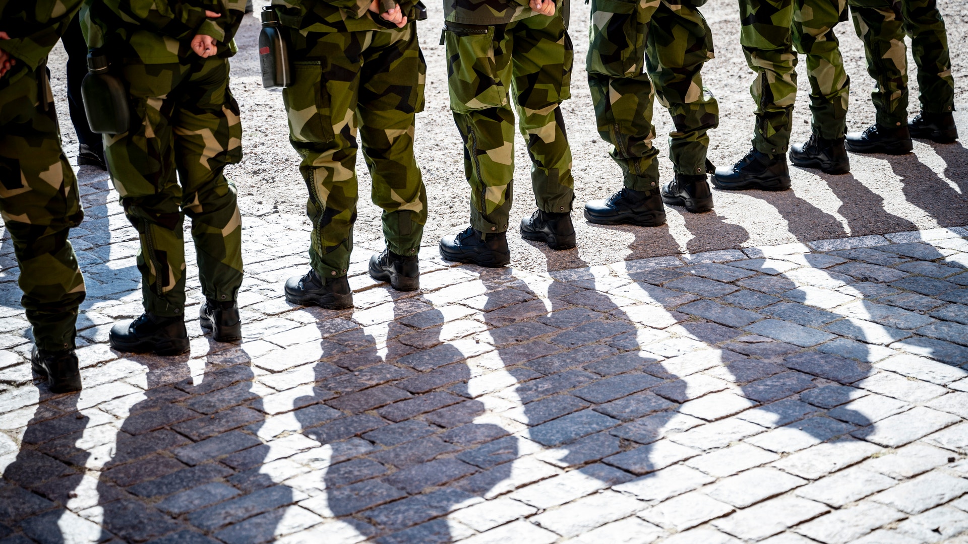 Soldater i kamouflageuniform står på rad på en stenlagd plats. Deras skuggor sträcker sig över stenarna.