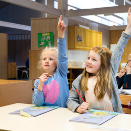 Elever i ett klassrum sitter vid bänkar och räcker upp händerna för att svara på frågor.