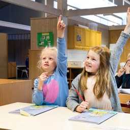 Elever i ett klassrum sitter vid bänkar och räcker upp händerna för att svara på frågor.