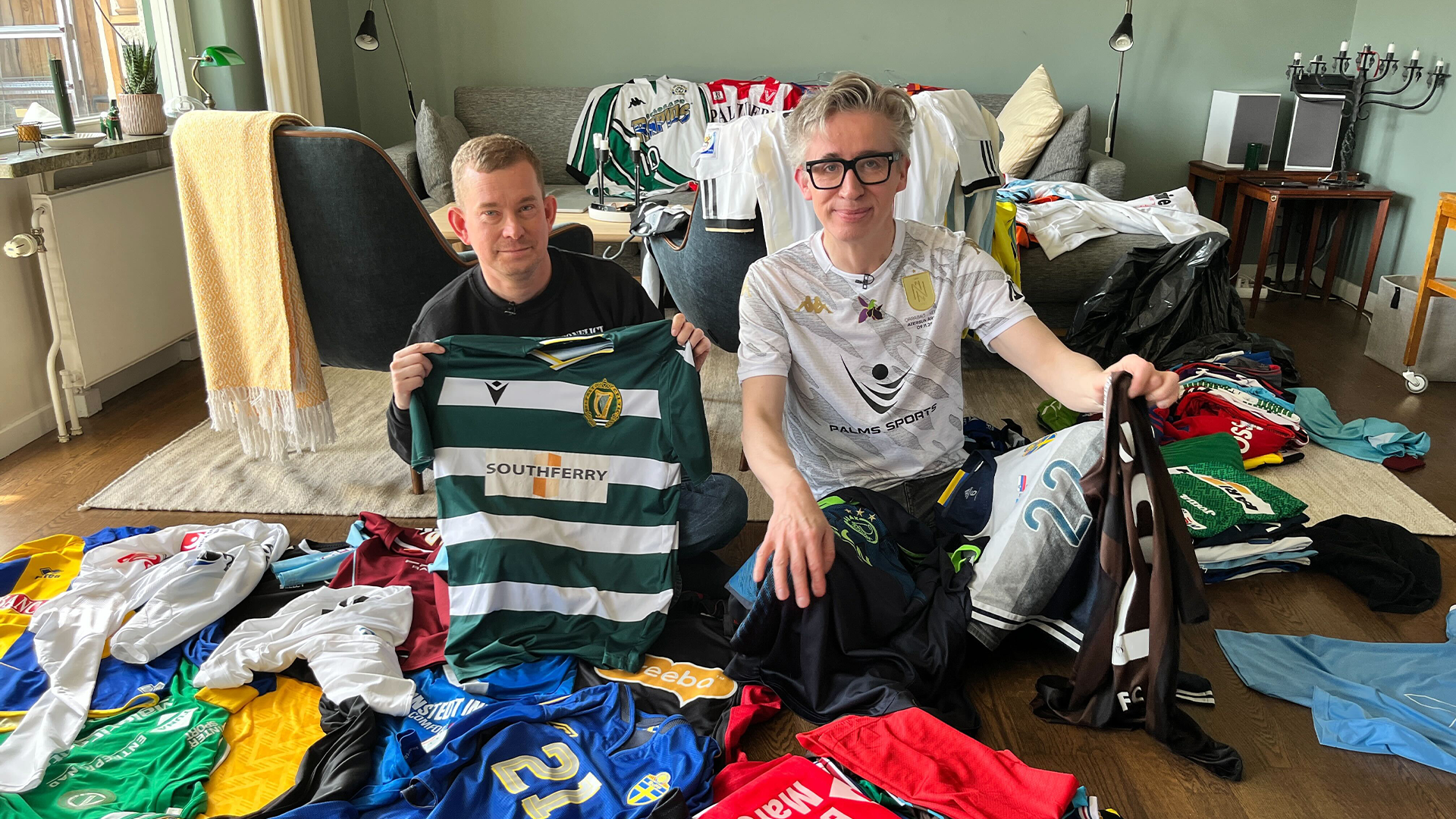 Två personer sitter på golvet omgivna av många färgglada klädesplagg. De håller upp en grön och vit randig sportdräkt.