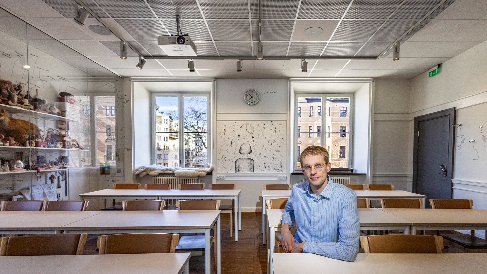 En man i blå skjorta sitter vid ett skrivbord i ett ljust klassrum eller kontor med fönster och bokhyllor i bakgrunden.