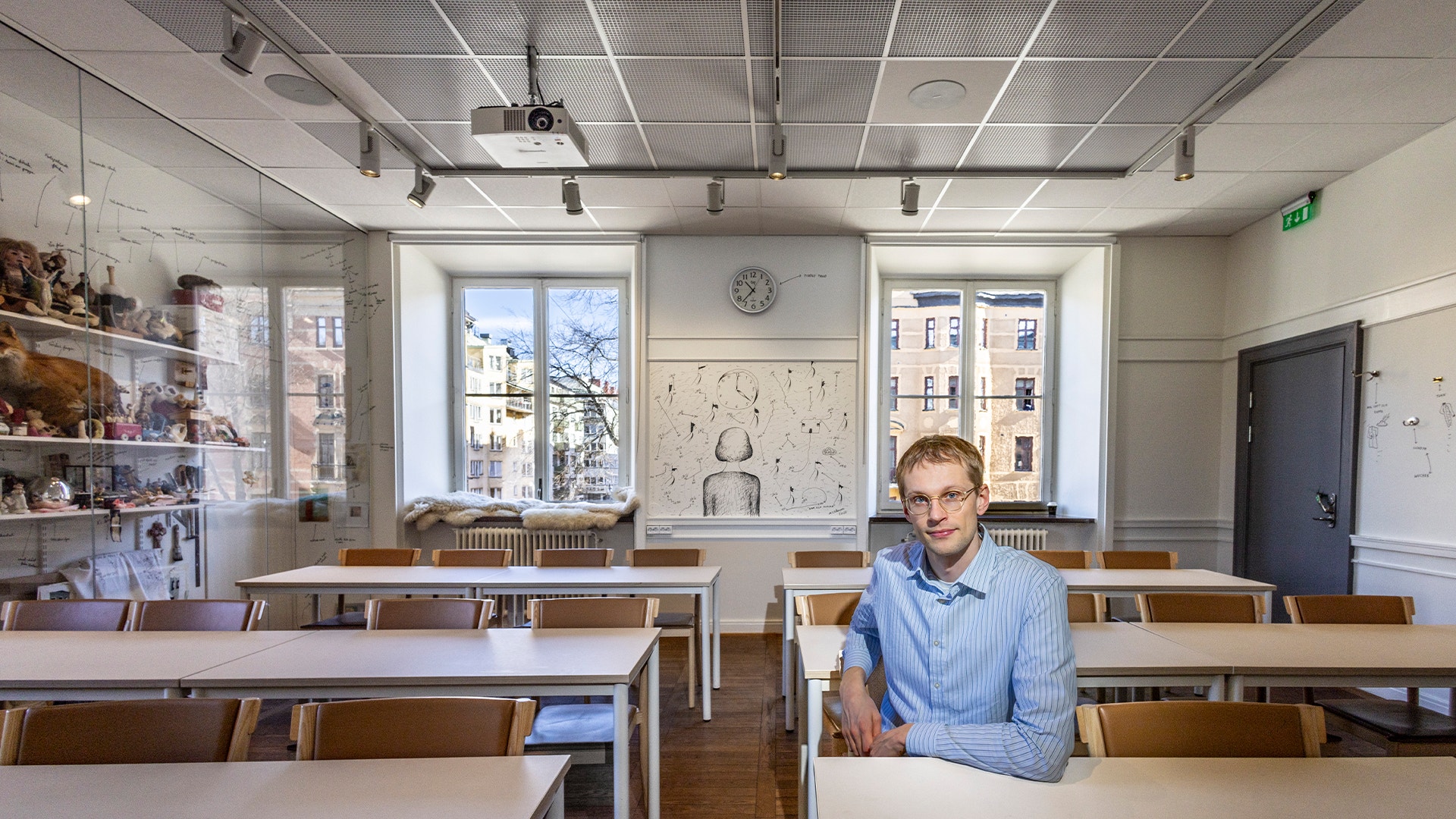 En man i blå skjorta sitter vid ett skrivbord i ett ljust klassrum eller kontor med fönster och bokhyllor i bakgrunden.