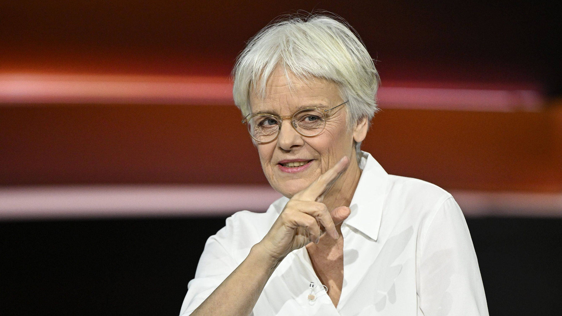 Porträttfoto av en  Ulrike Herrmann med kort vitt hår och glasögon, klädd i vit skjorta. Hon poserar med handen mot ansiktet i en thoughtful hållning mot en varm orange och röd bakgrund.
