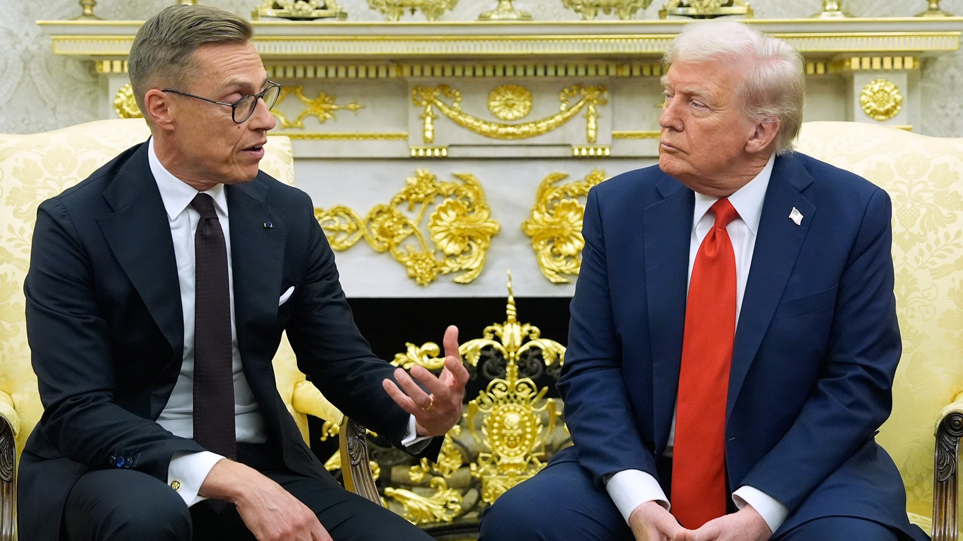 Två män i formell affärsdräkt sitter och samtalar i ett prydligt dekorerat rum med guldiga ornament.