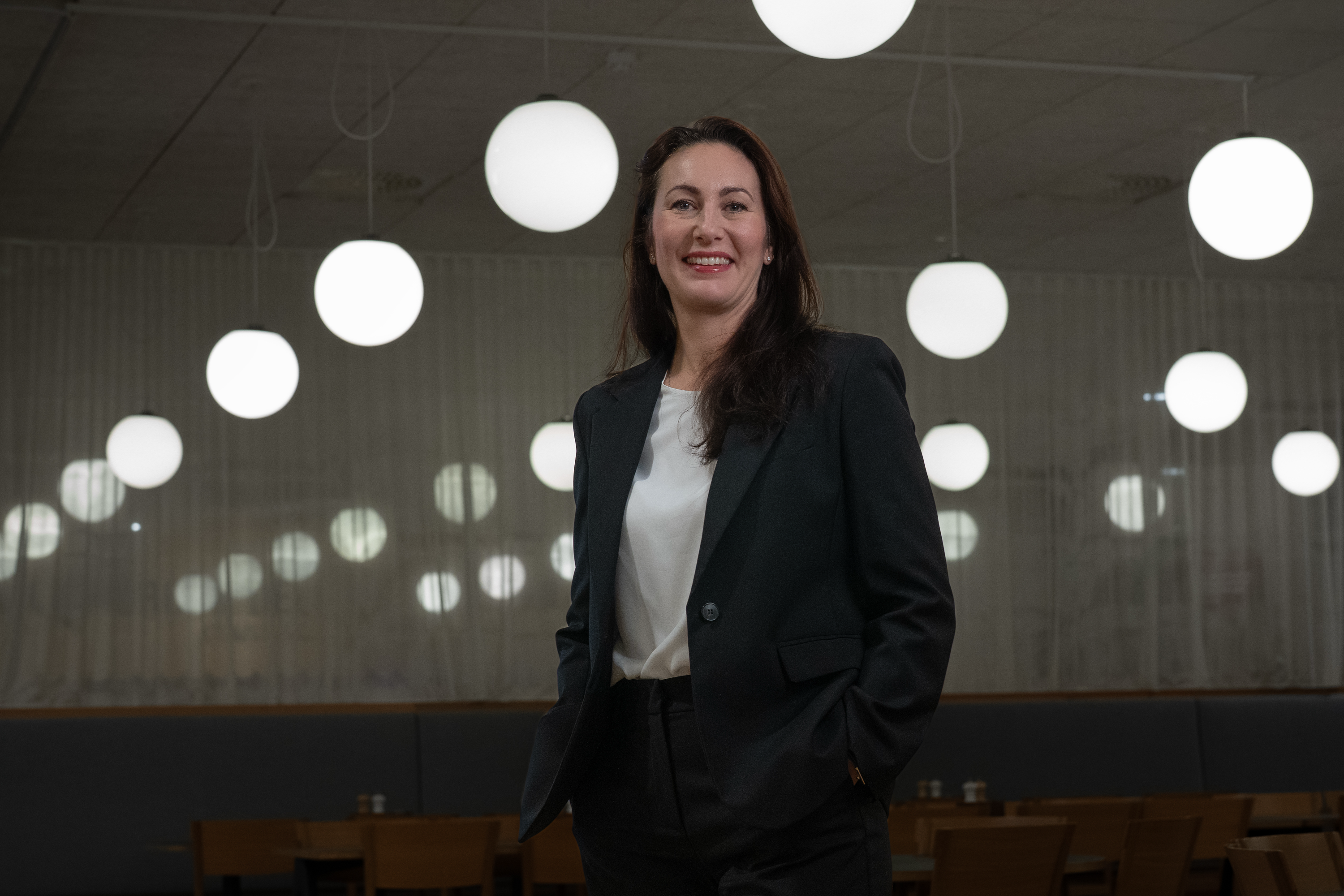 Leende kvinna med långt mörkt hår, klädd i svart kavaj och vit blus, står med handen i fickan i en restaurang med runda, vita hänglampor.