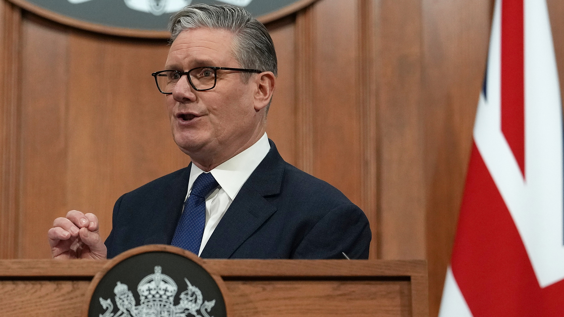 En man i kostym och glasögon talar vid ett officiellt podium med brittisk regeringsemblem. En brittisk flagga syns i bakgrunden.