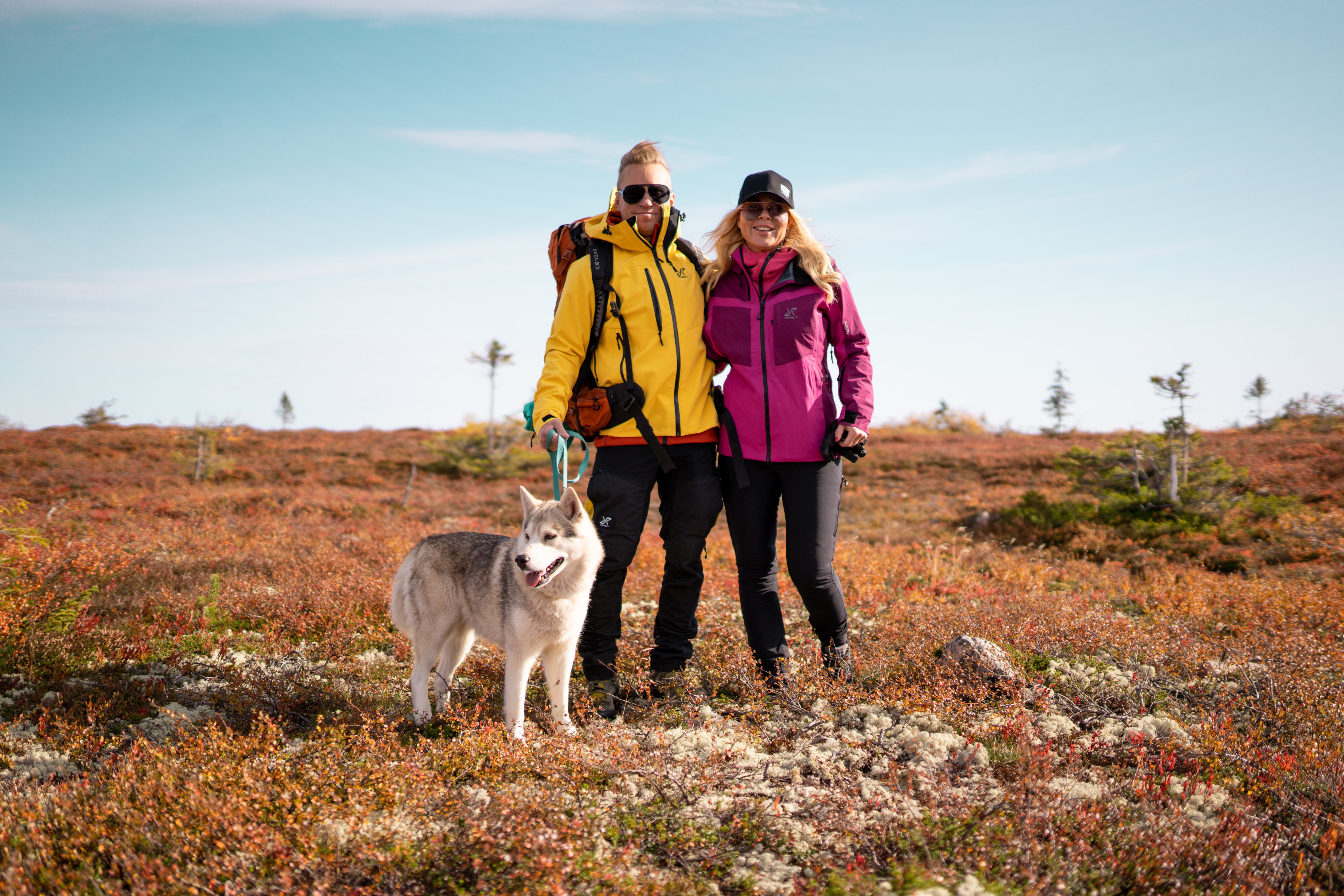 Två personer i färgglada jackor står tillsammans med en husky i ett öppet, kuperat landskap under blå himmel.