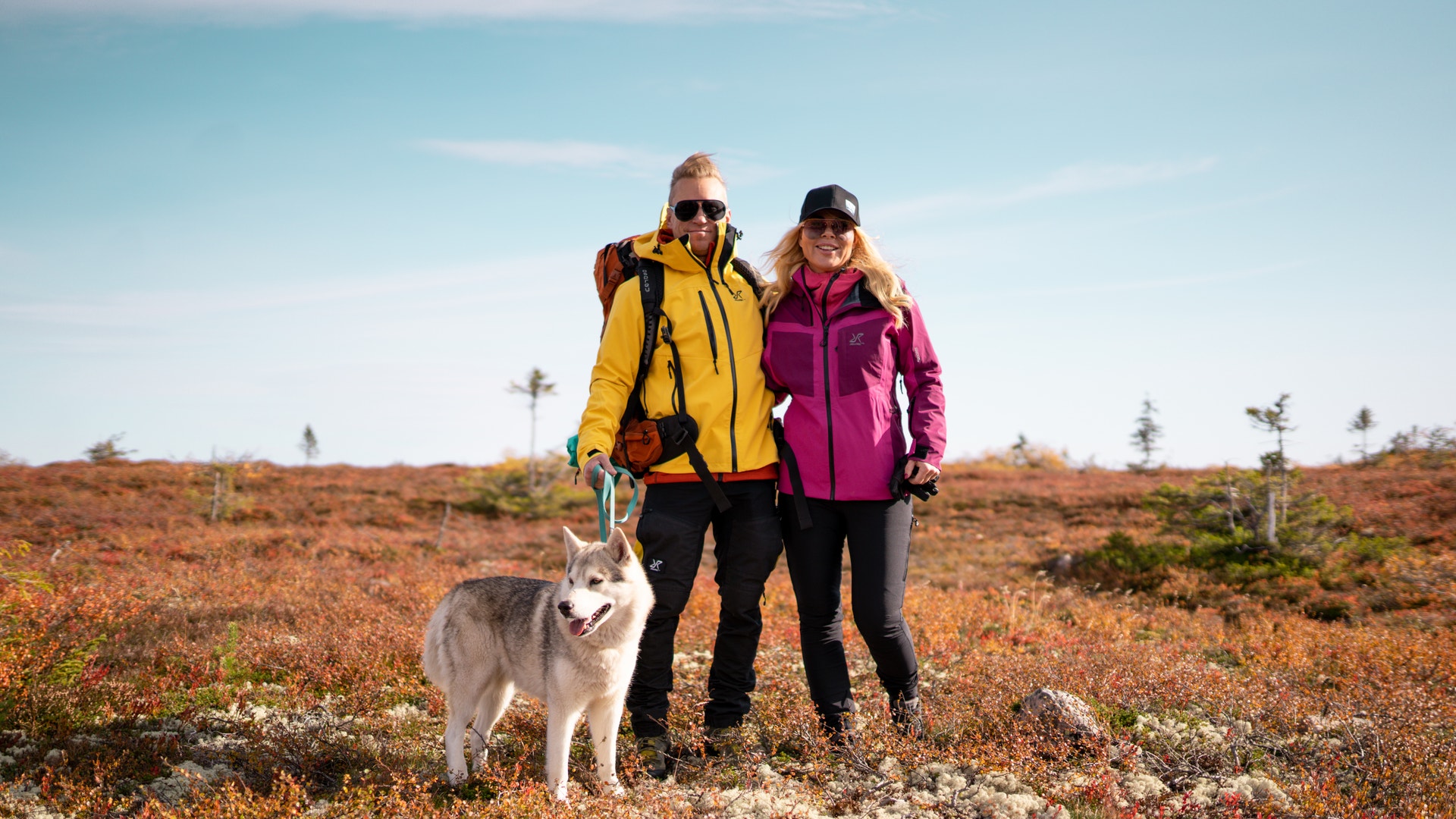 Två personer i färgglada jackor står tillsammans med en husky i ett öppet, kuperat landskap under blå himmel.