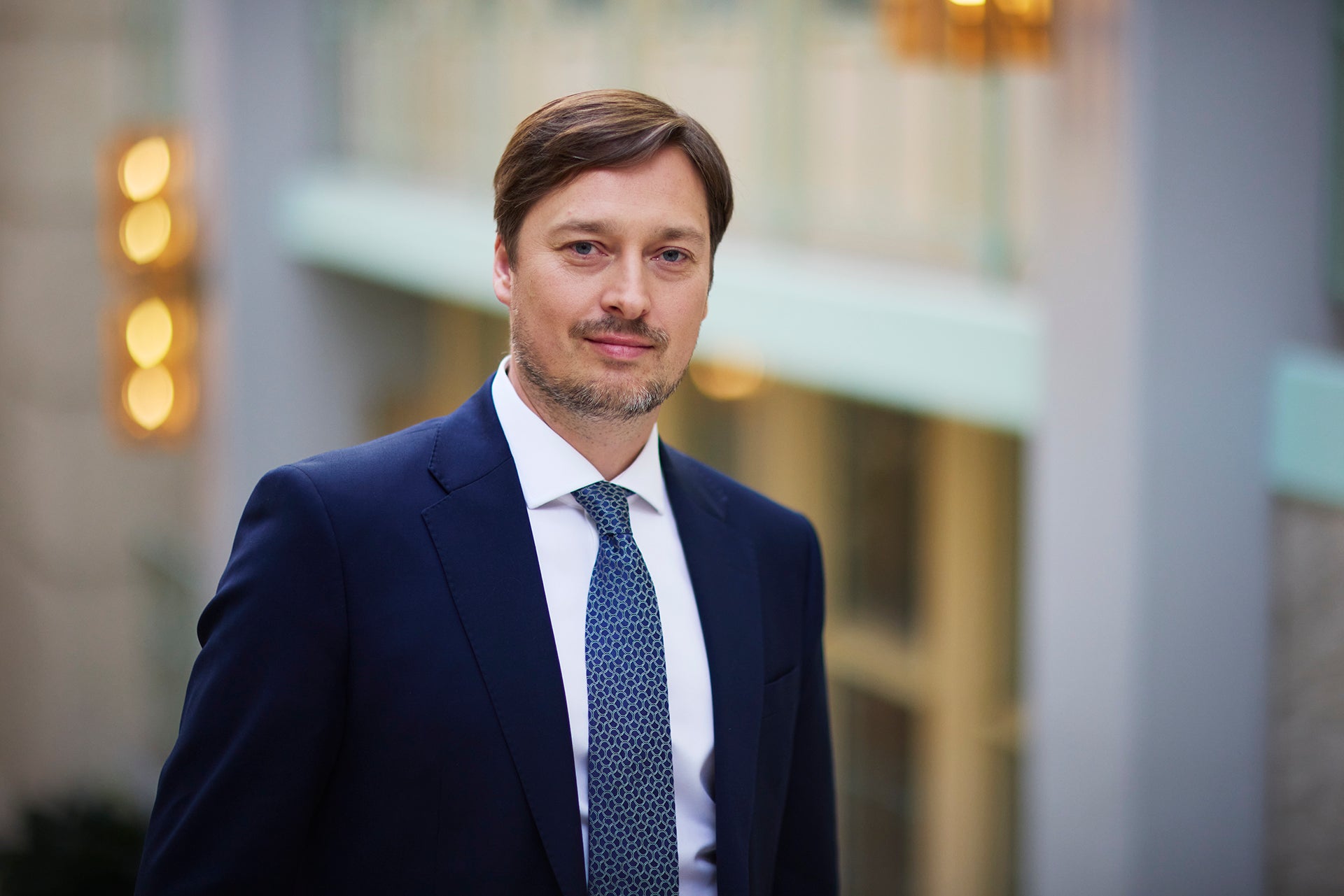 Porträtt av en man i mörkblå kostym och slips framför en suddig stadsmiljö.