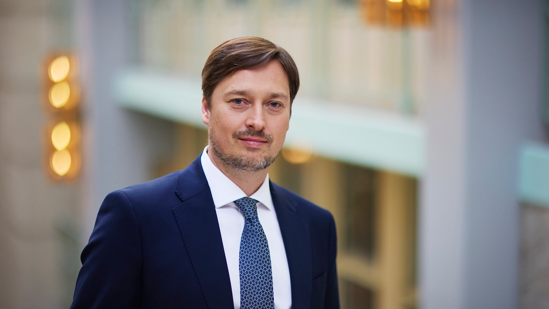 Porträtt av en man i mörkblå kostym och slips framför en suddig stadsmiljö.