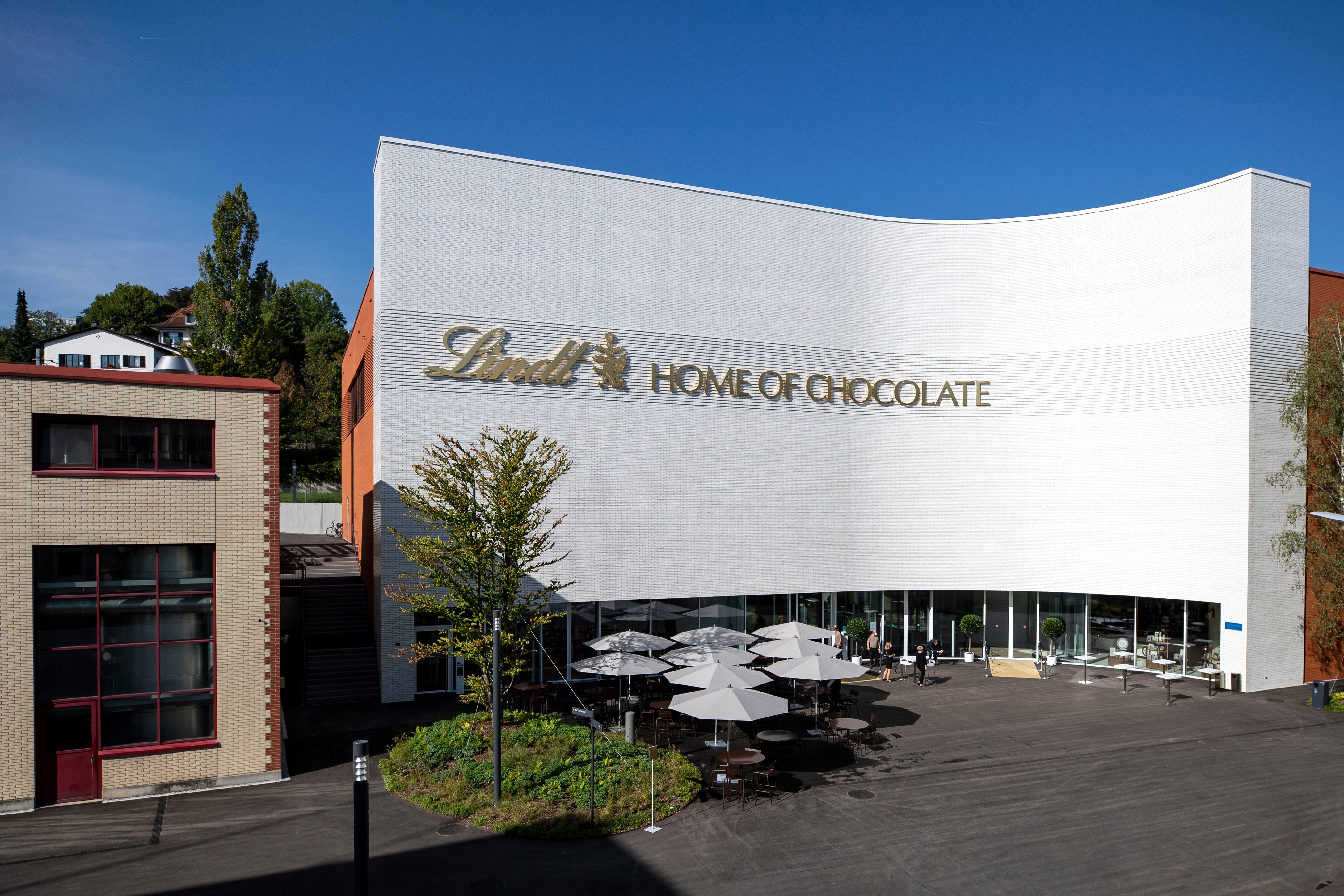 En vit, böljande byggnad med texten "Lindt Home of Chocolate" på fasaden, omgiven av parasoller och grönska.