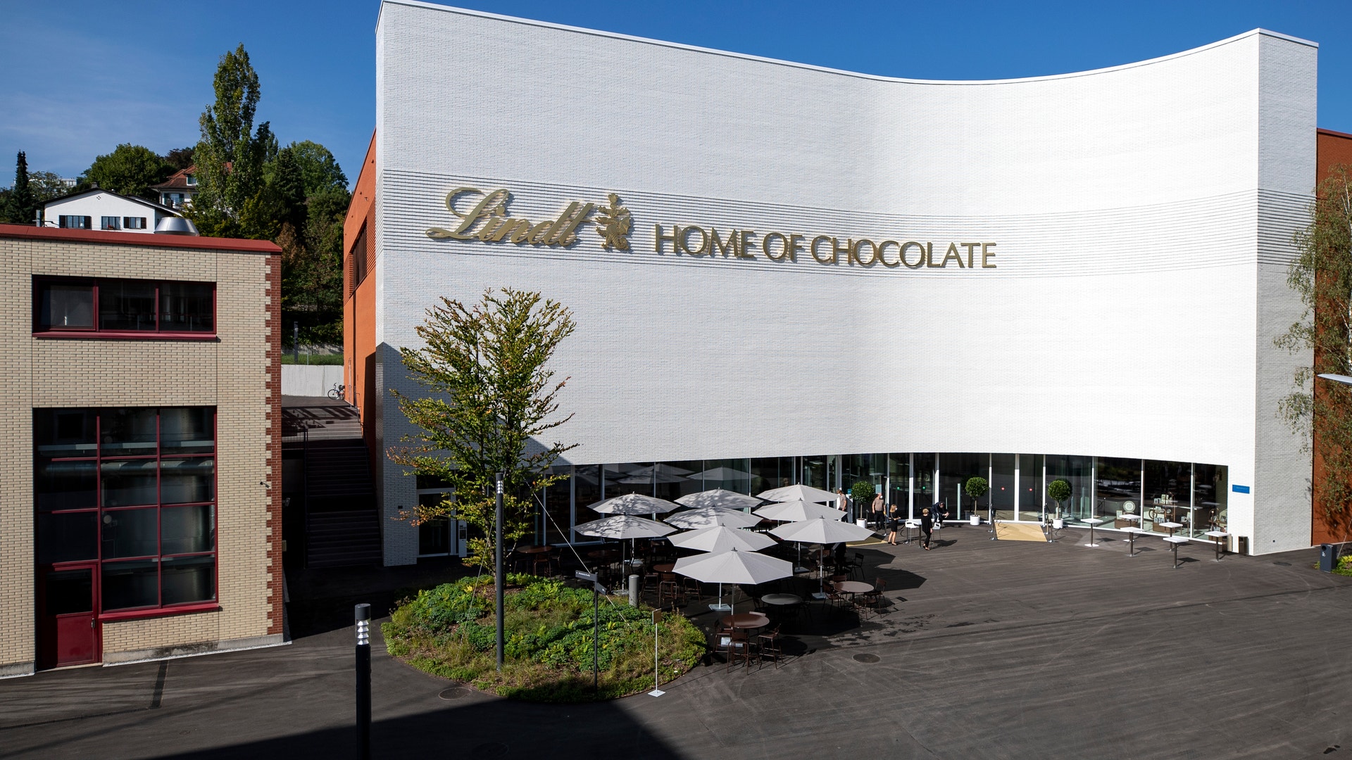En vit, böljande byggnad med texten "Lindt Home of Chocolate" på fasaden, omgiven av parasoller och grönska.