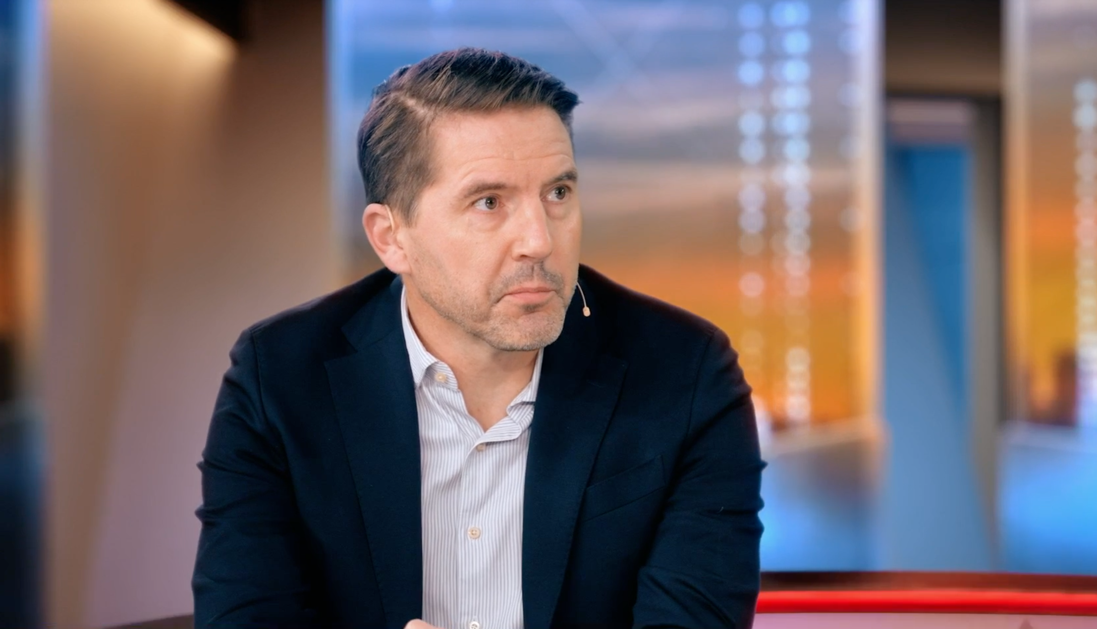 En man i mörk kavaj och randig skjorta sitter i en tv-studio och tittar åt sidan.