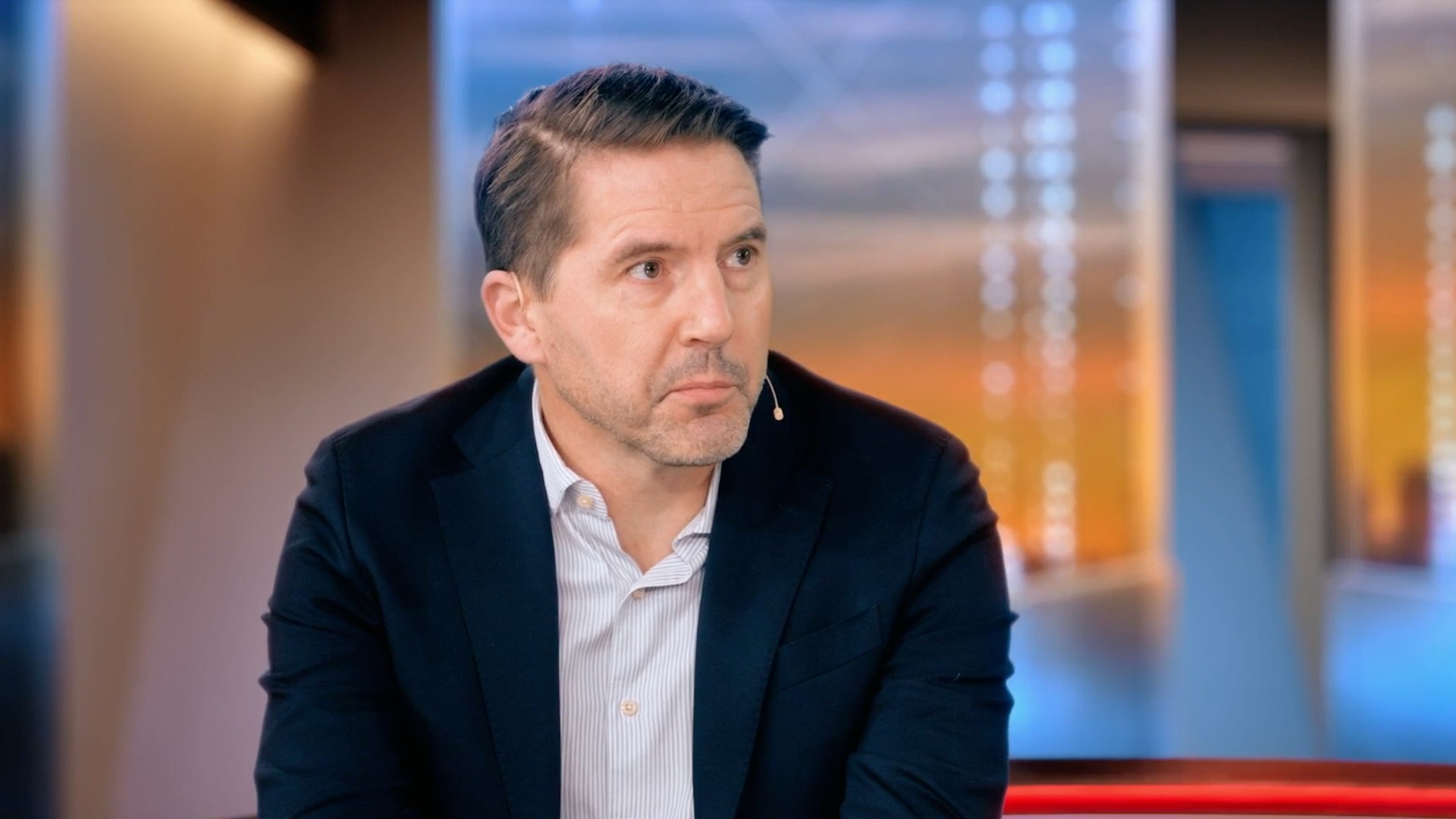 En man i mörk kavaj och randig skjorta sitter i en tv-studio och tittar åt sidan.