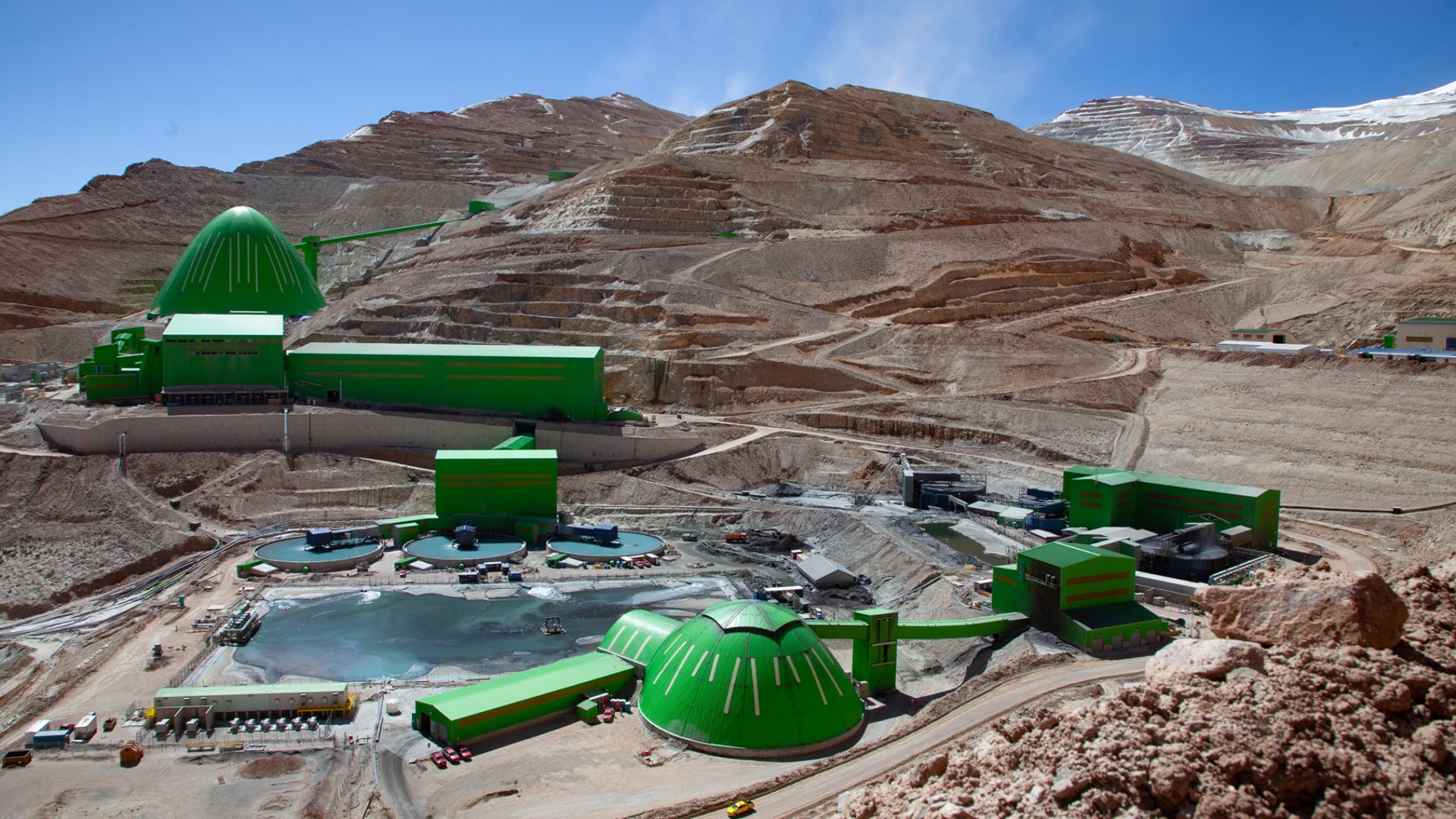 Luftfoto av en gruvdrift med grönt gruvutrustning och behandlingsanläggningar placerade i ett kalt, bergigt ökenlandskap. Flera stora gröna metallkonstruktioner och behållare är synliga tillsammans med gruvarbetsplatser och vägar i det kala, steniga terrängen.