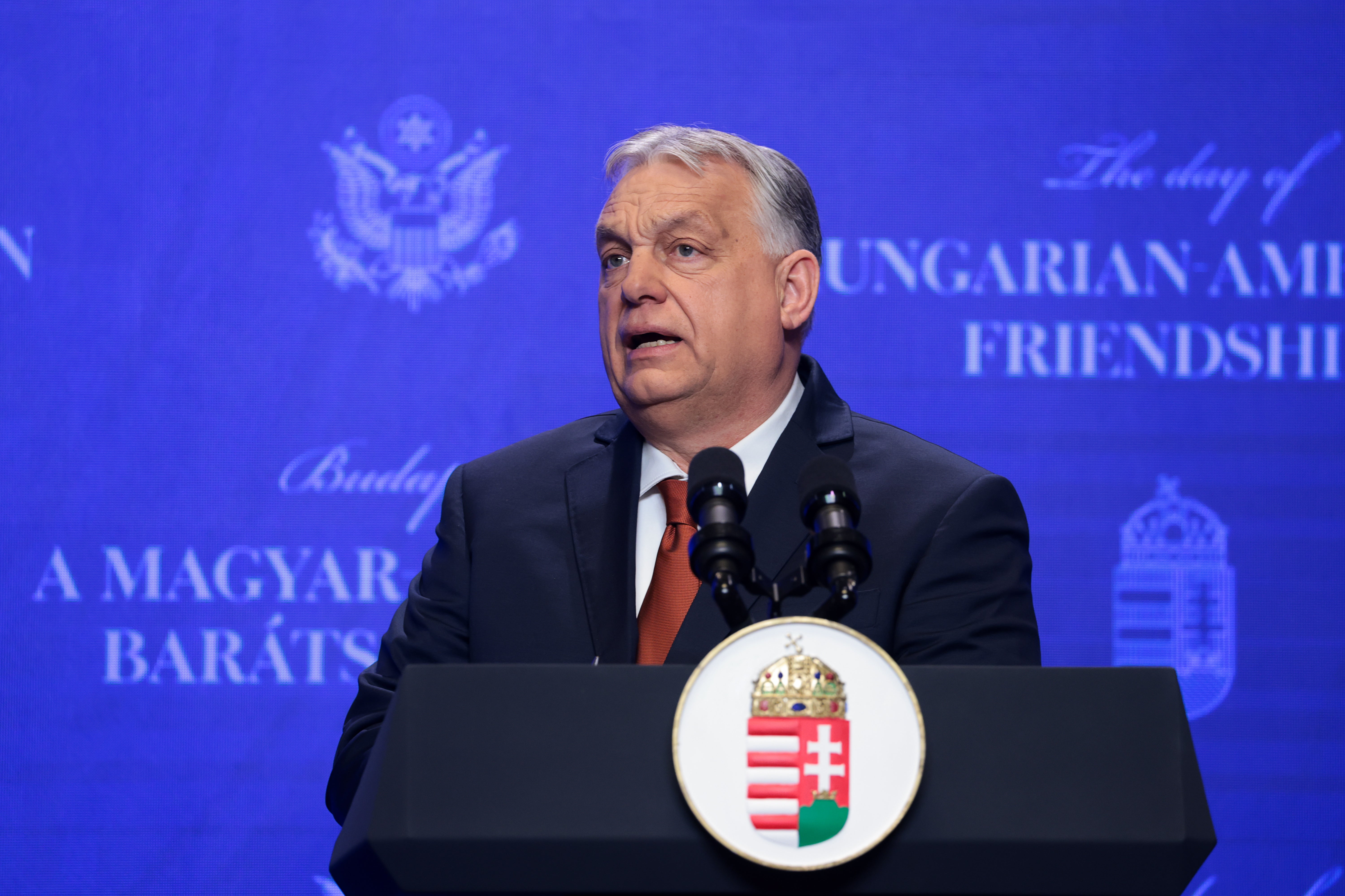 Man i mörk kostym och röd slips talar vid ett podium med Ungerns vapensköld framför en blå bakgrund.