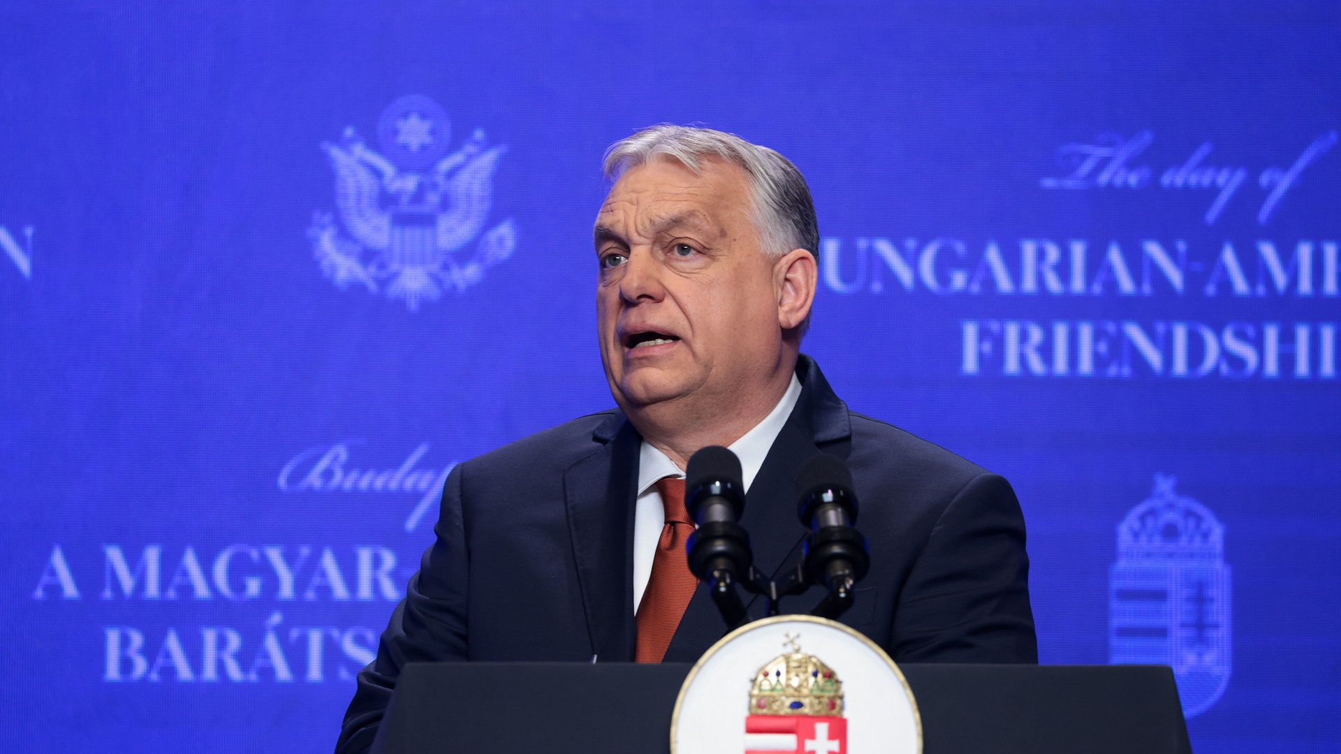 Man i mörk kostym och röd slips talar vid ett podium med Ungerns vapensköld framför en blå bakgrund.