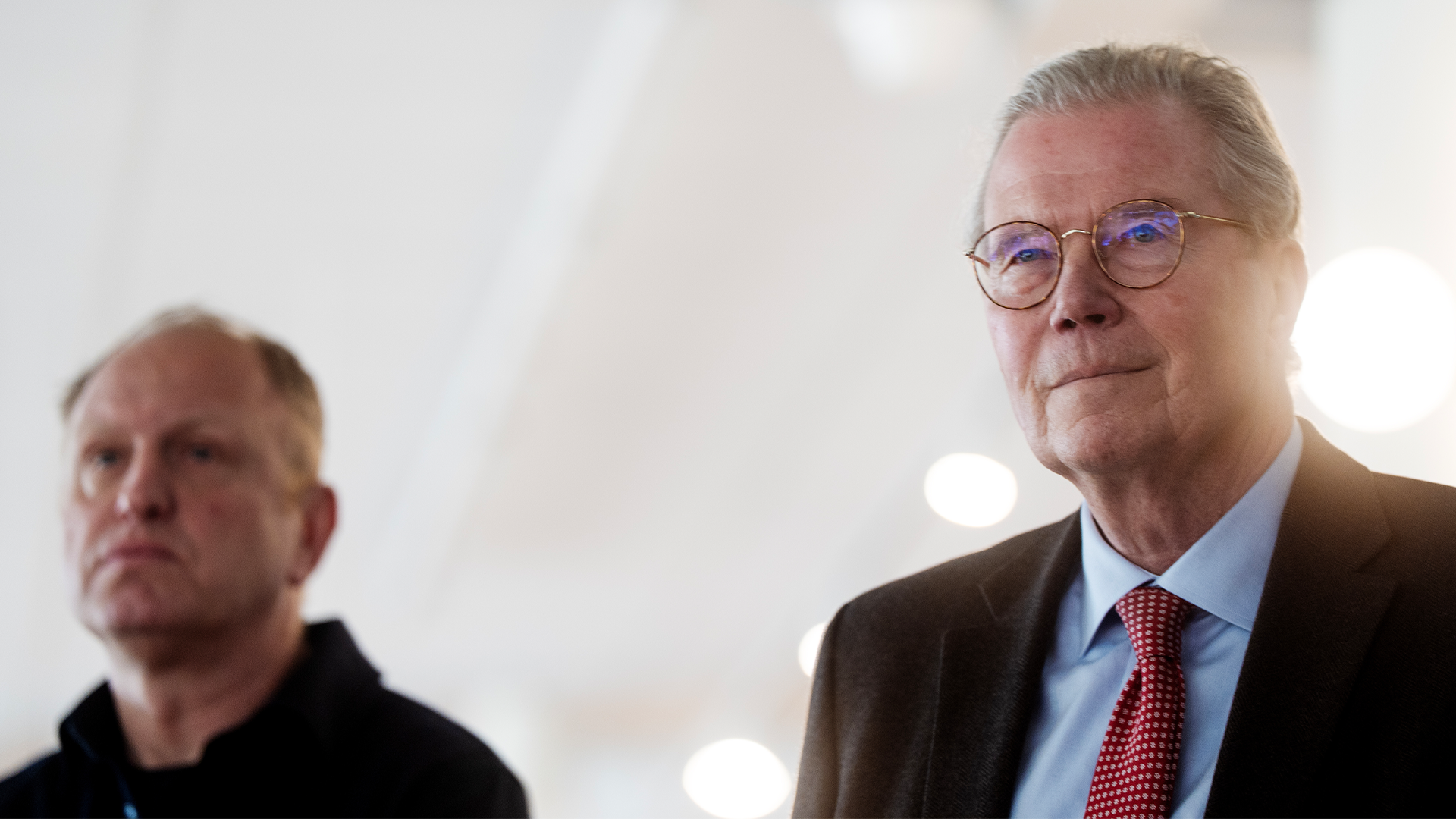 Två män i professionell miljö. Mannen i förgrunden bär glasögon, blå skjorta och mörkbrun kavaj. En annan man syns suddig i bakgrunden.