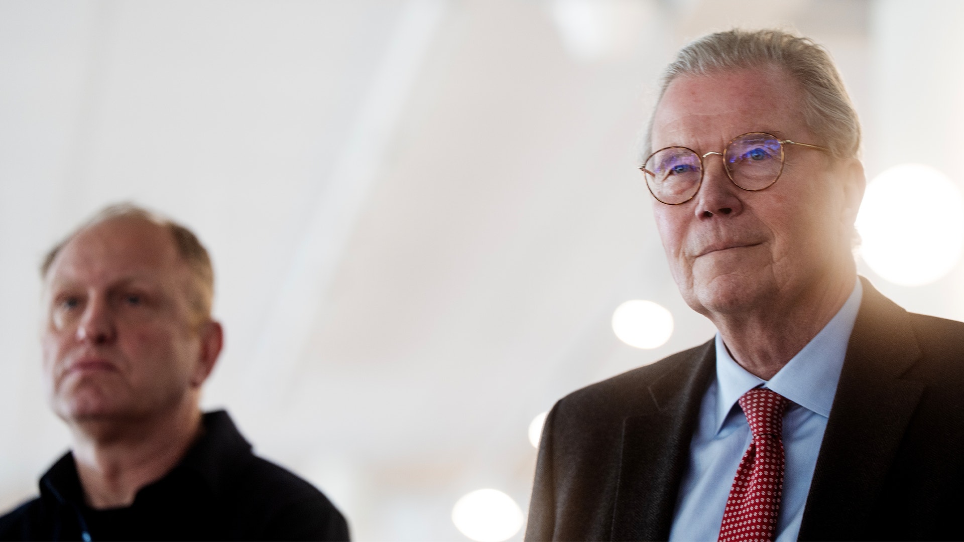 Två män i professionell miljö. Mannen i förgrunden bär glasögon, blå skjorta och mörkbrun kavaj. En annan man syns suddig i bakgrunden.