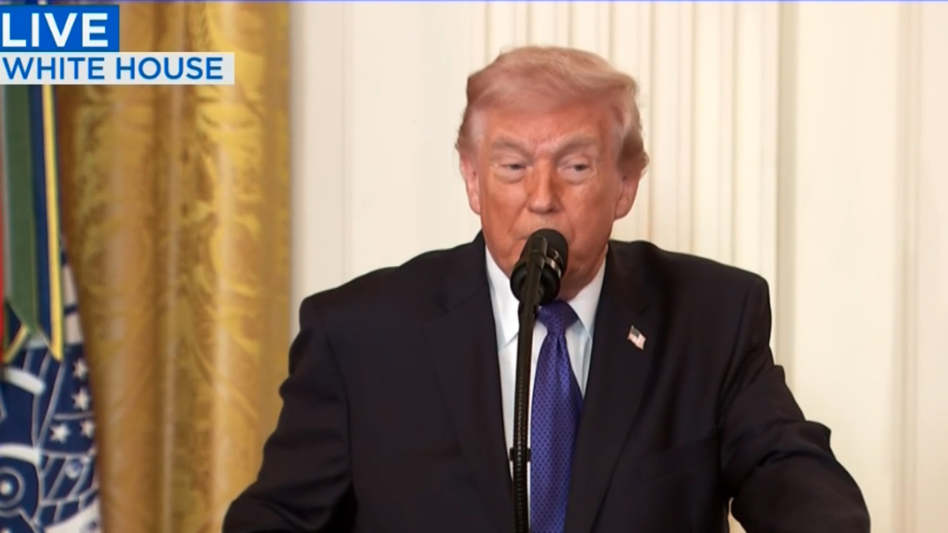 Man i mörk kostym och blå slips talar vid mikrofon under en direkt sändning från Vita huset.