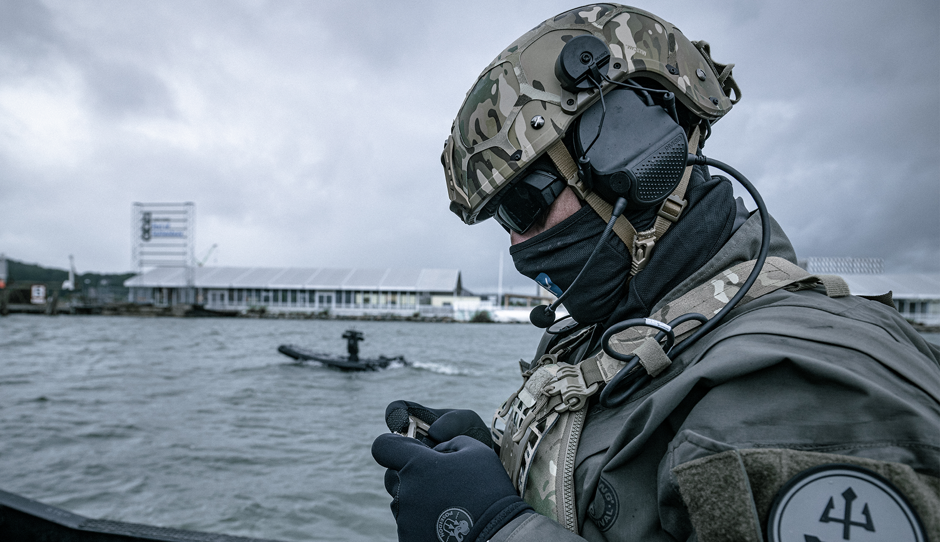 En kamouflageklädd soldat med hjälm och headset står på en båt vid en hamn, med en mindre båt synlig i vattnet bakom.